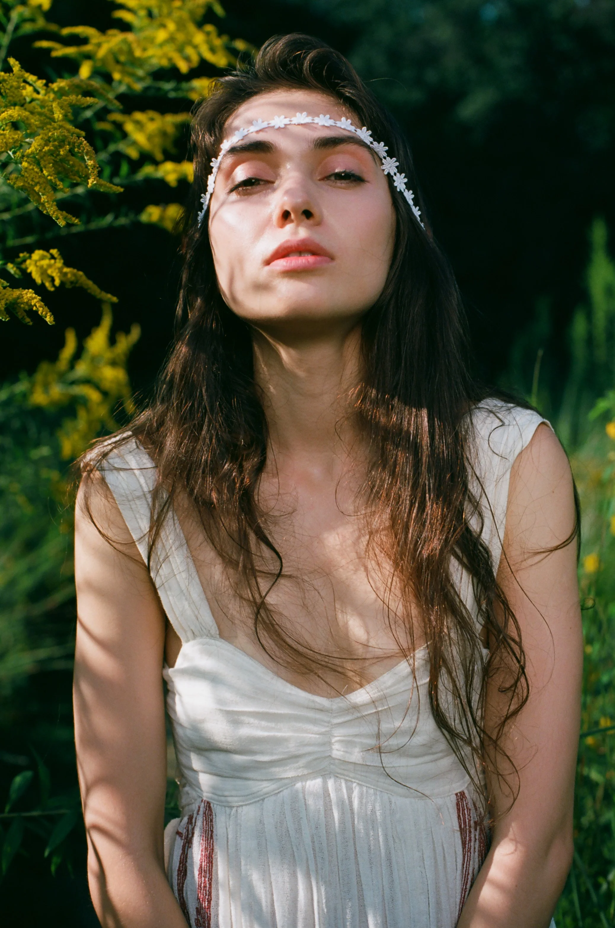 A young woman with long, dark wavy hair and light skin wearing a white dress and a white floral headband, standing outdoors in a lush green setting with yellow flowers and dark greenery in the background.