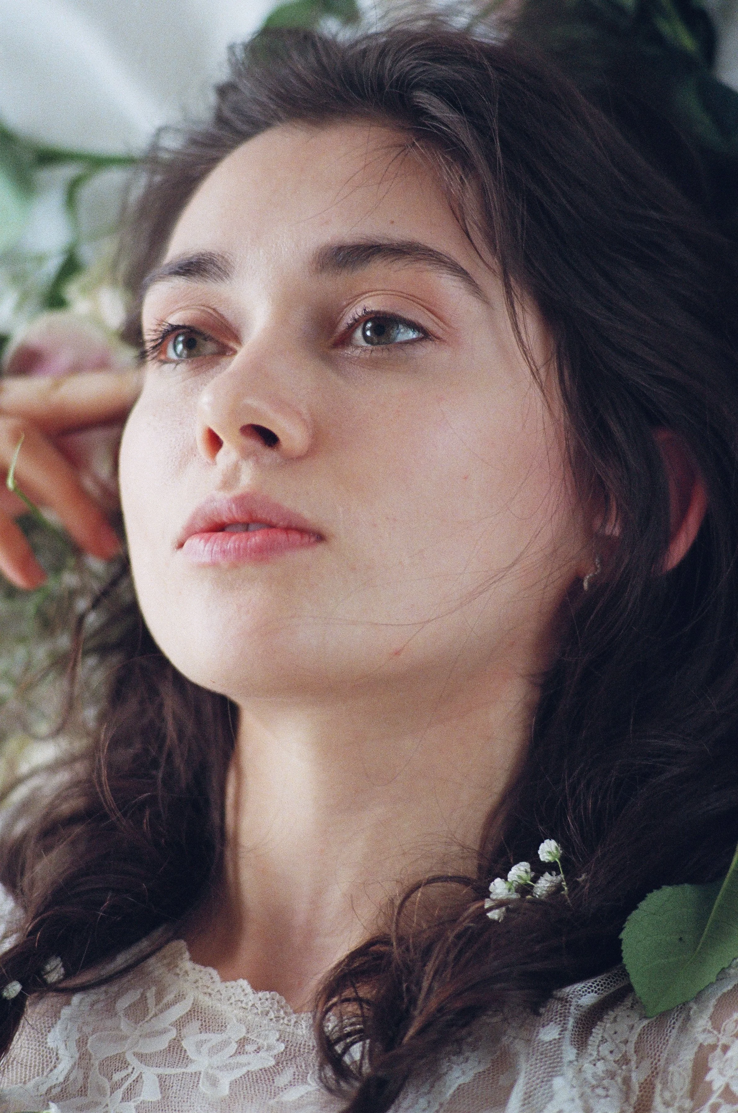 Close-up of a young woman with long dark hair, lying down among green leaves and flowers, wearing a lace top, looking thoughtful.