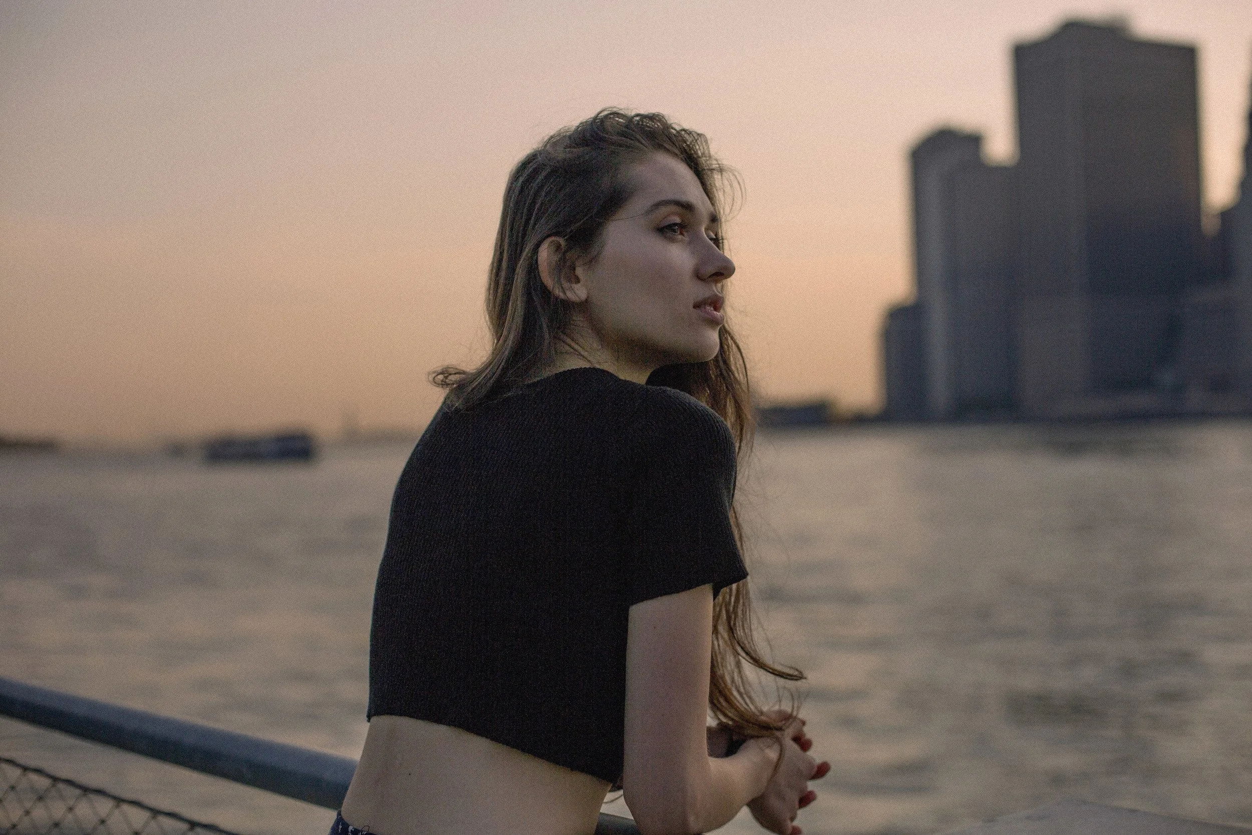 A woman with long brown hair and a black crop top standing by a body of water during sunset, with city buildings in the background.