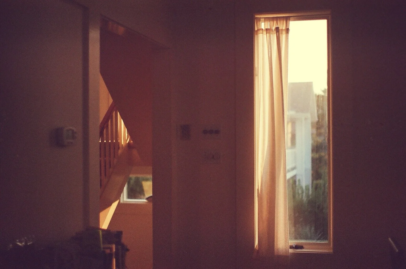 Indoor scene with a room illuminated by sunlight through a large window with beige curtains, showing a partial view of a staircase and exterior buildings.