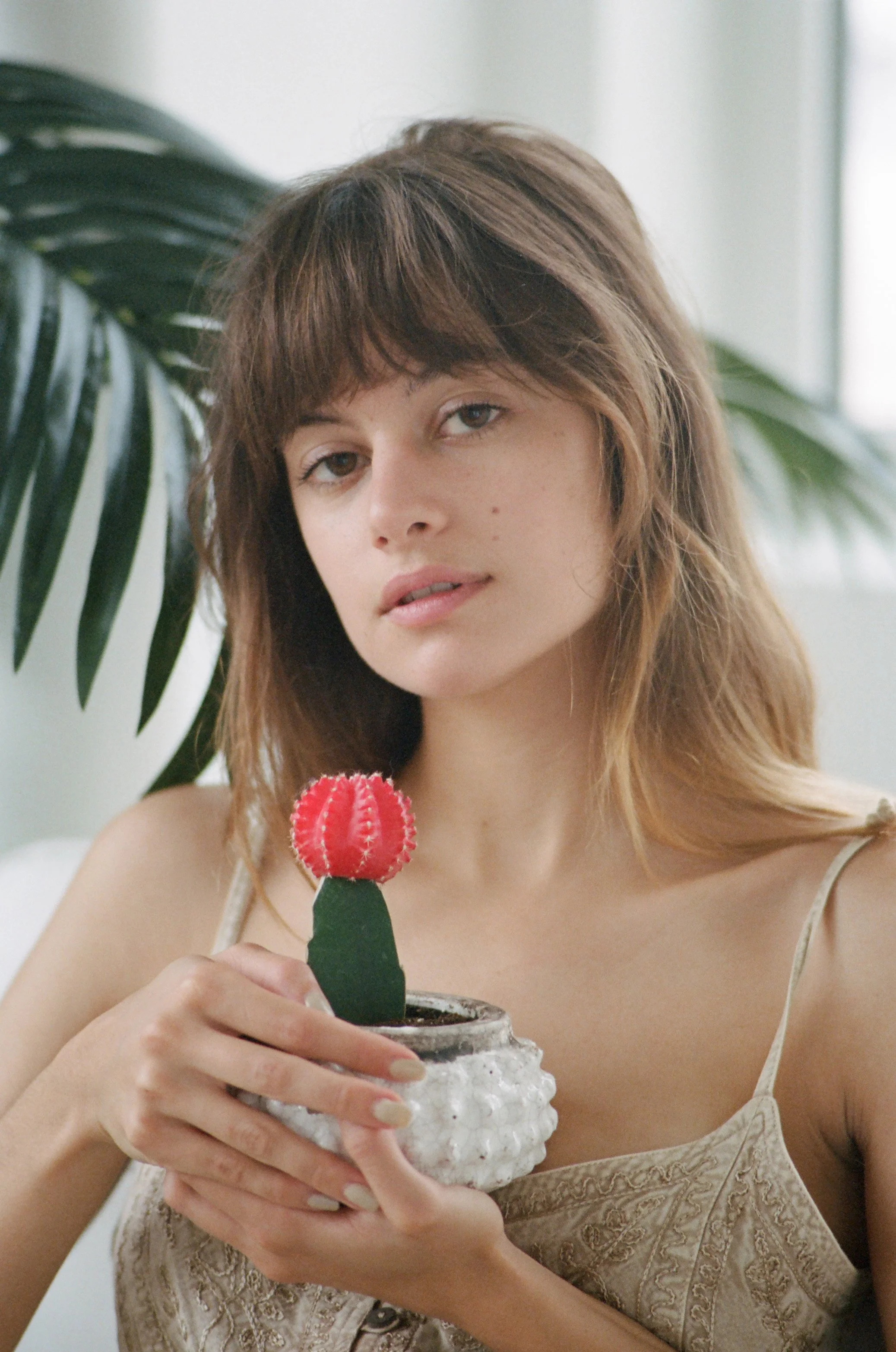 A young woman with brown hair and blue eyes holding a potted cactus shaped like a red flower, indoors with green tropical plant leaves in the background.