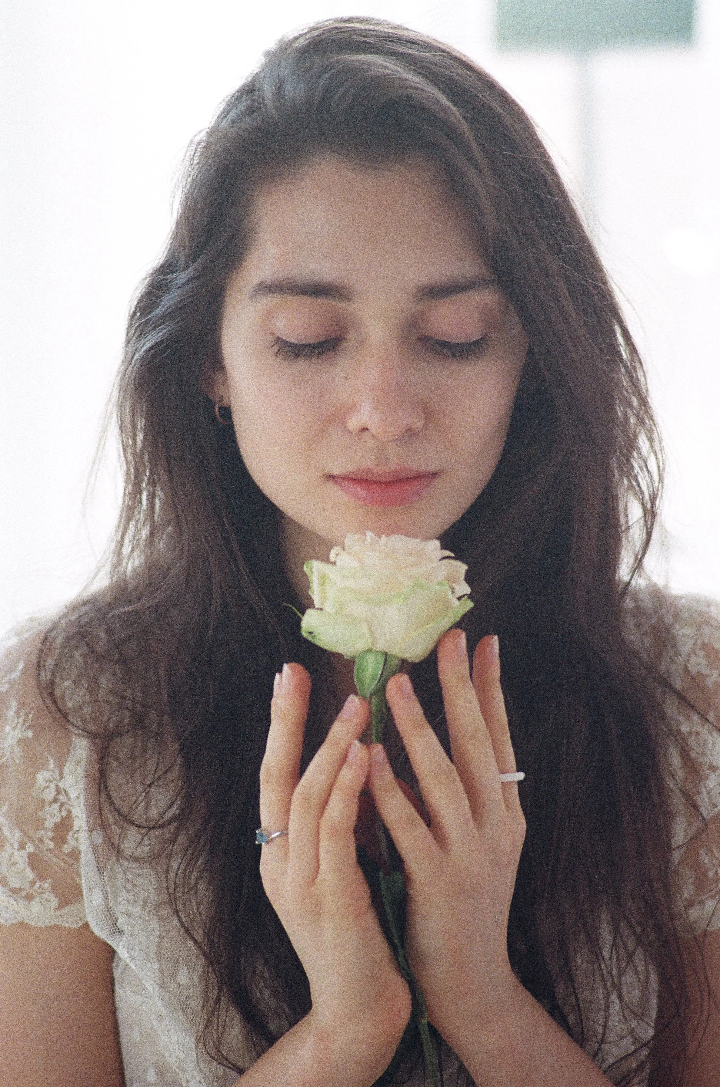 A young woman with dark hair, closed eyes, and earrings, holding a white rose close to her face.