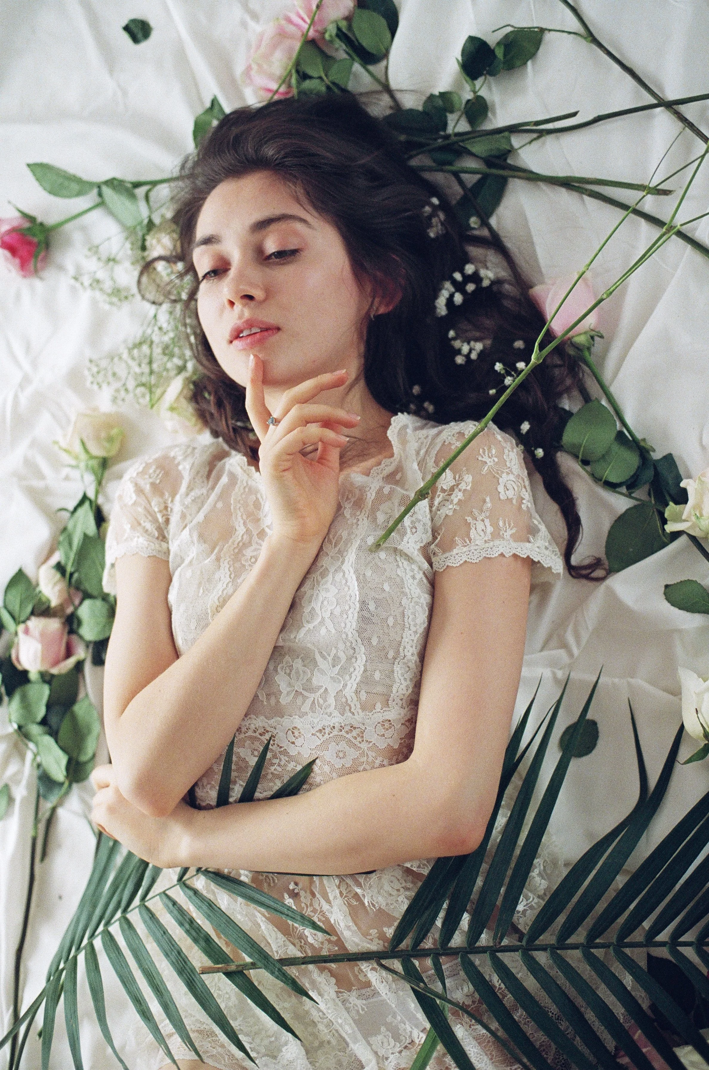 A woman lying on a bed surrounded by flowers, wearing a white lace dress, with dark hair and a thoughtful expression.