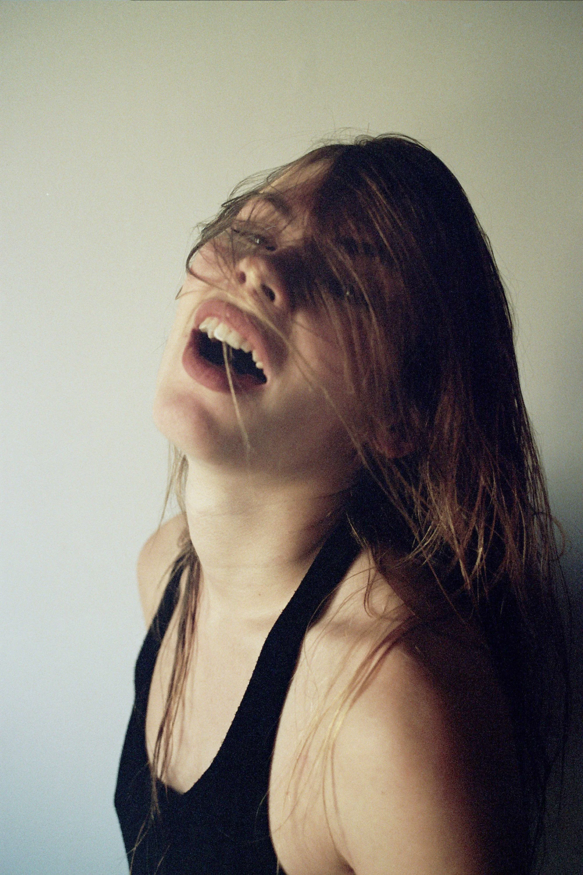 A woman with long brown hair and light skin, tilting her head back with her mouth open, wearing a black sleeveless top.