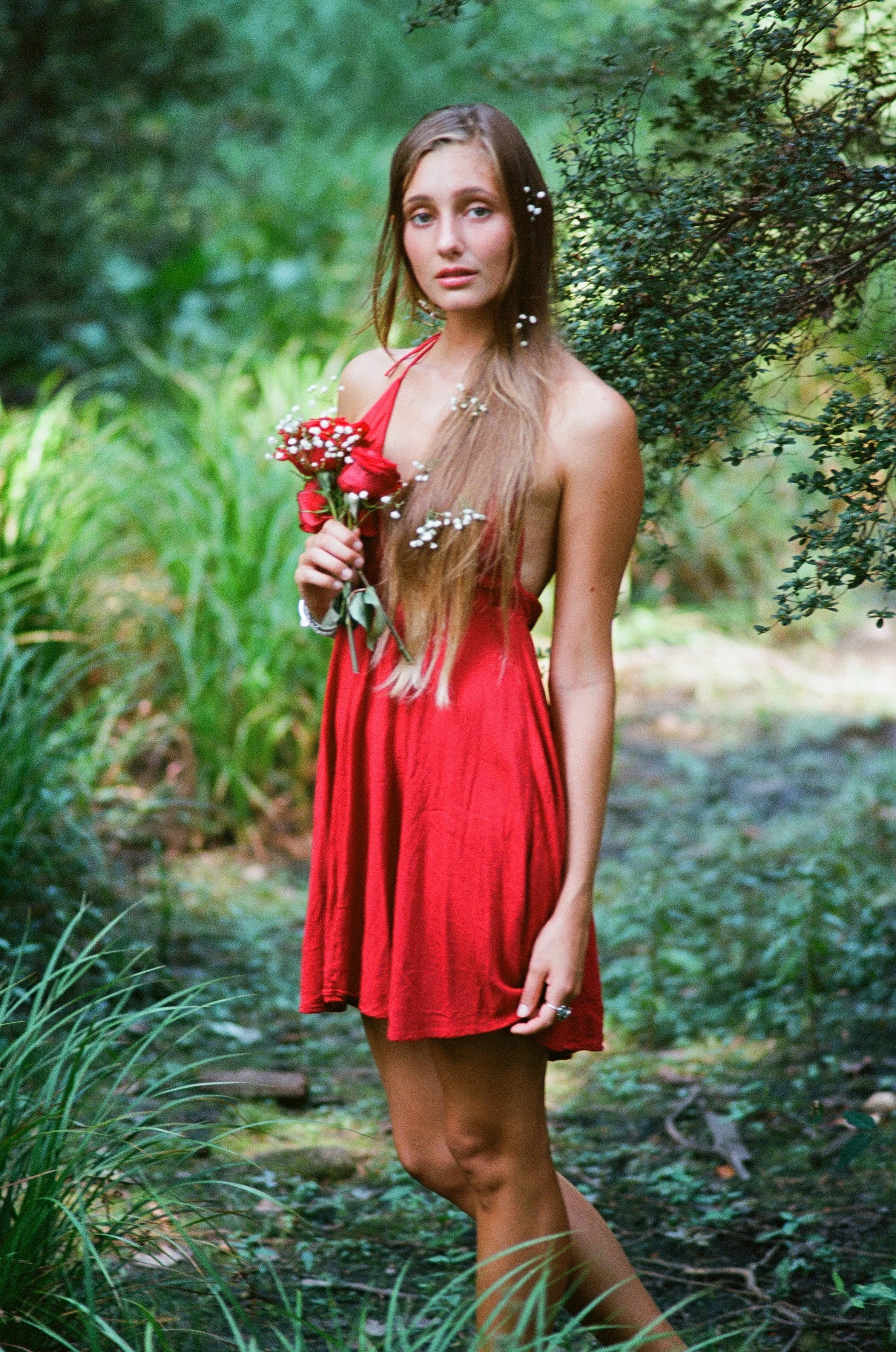 Young woman with long blonde hair wearing a red dress holding a bouquet of red roses and white flowers in a lush green outdoor setting.