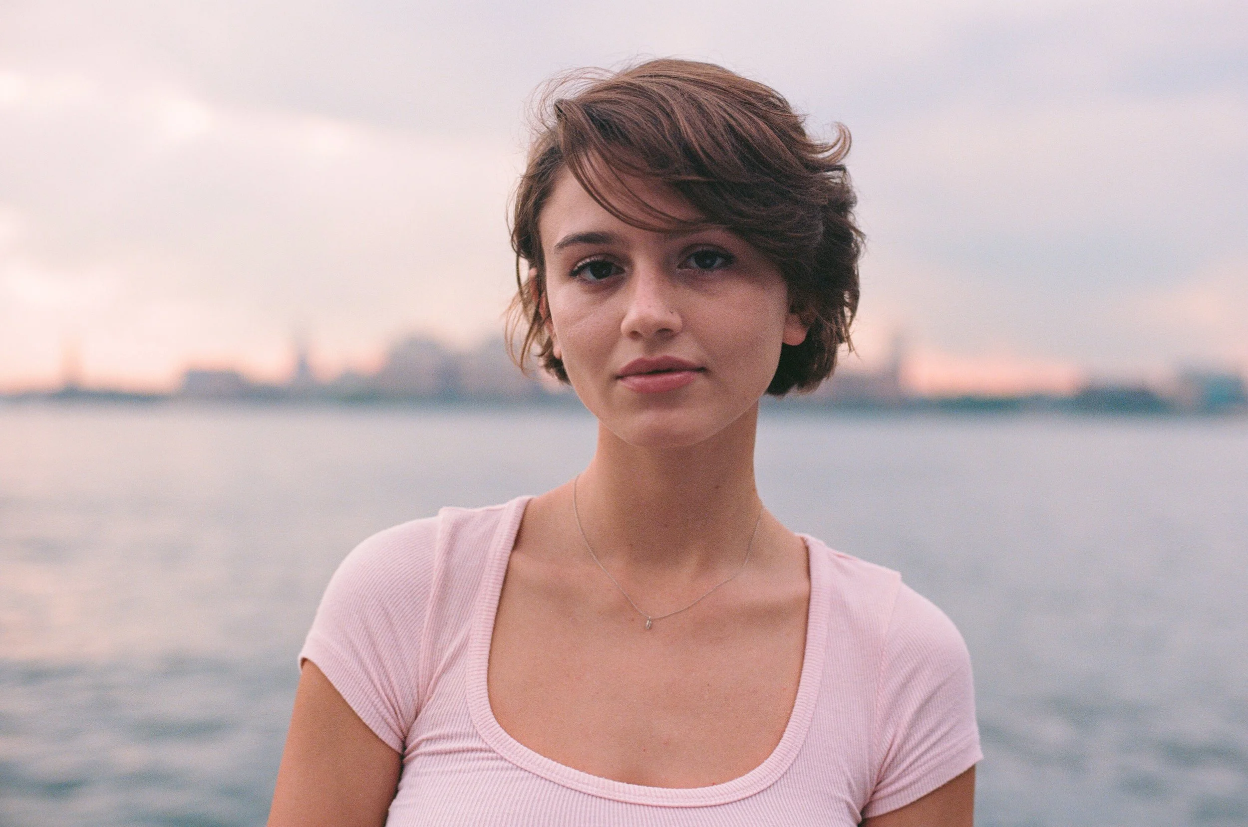 A young woman with short brown hair, wearing a light pink t-shirt and a delicate necklace, standing outdoors near a body of water with a city skyline in the background, during an overcast day.