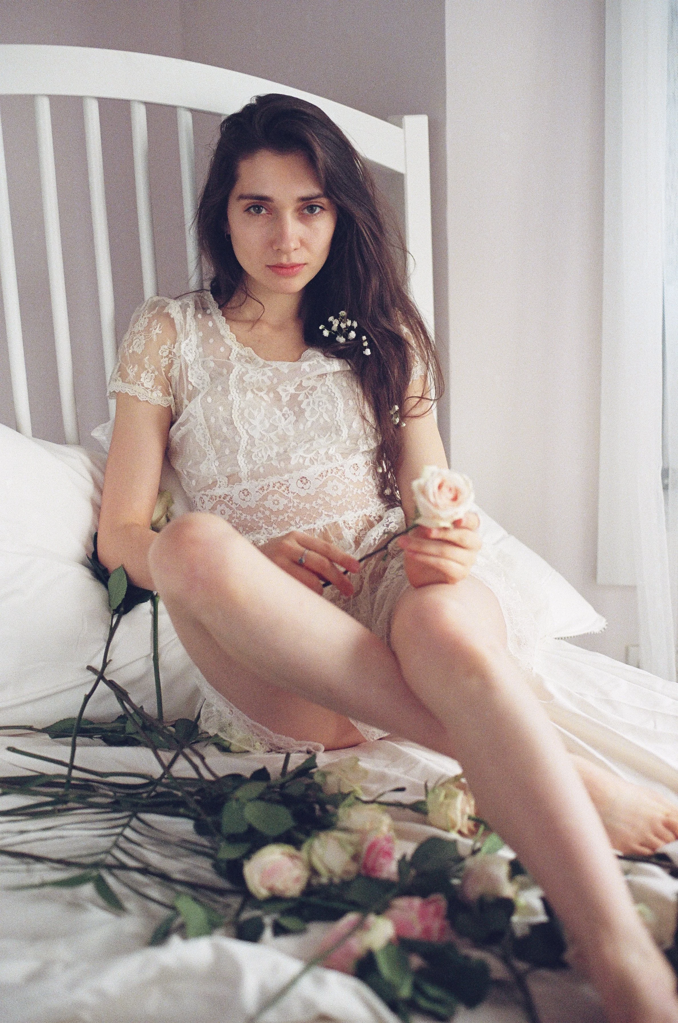 A young woman with long dark hair sitting on a bed, wearing a white lace dress, holding a pink rose, surrounded by fallen roses and greenery.