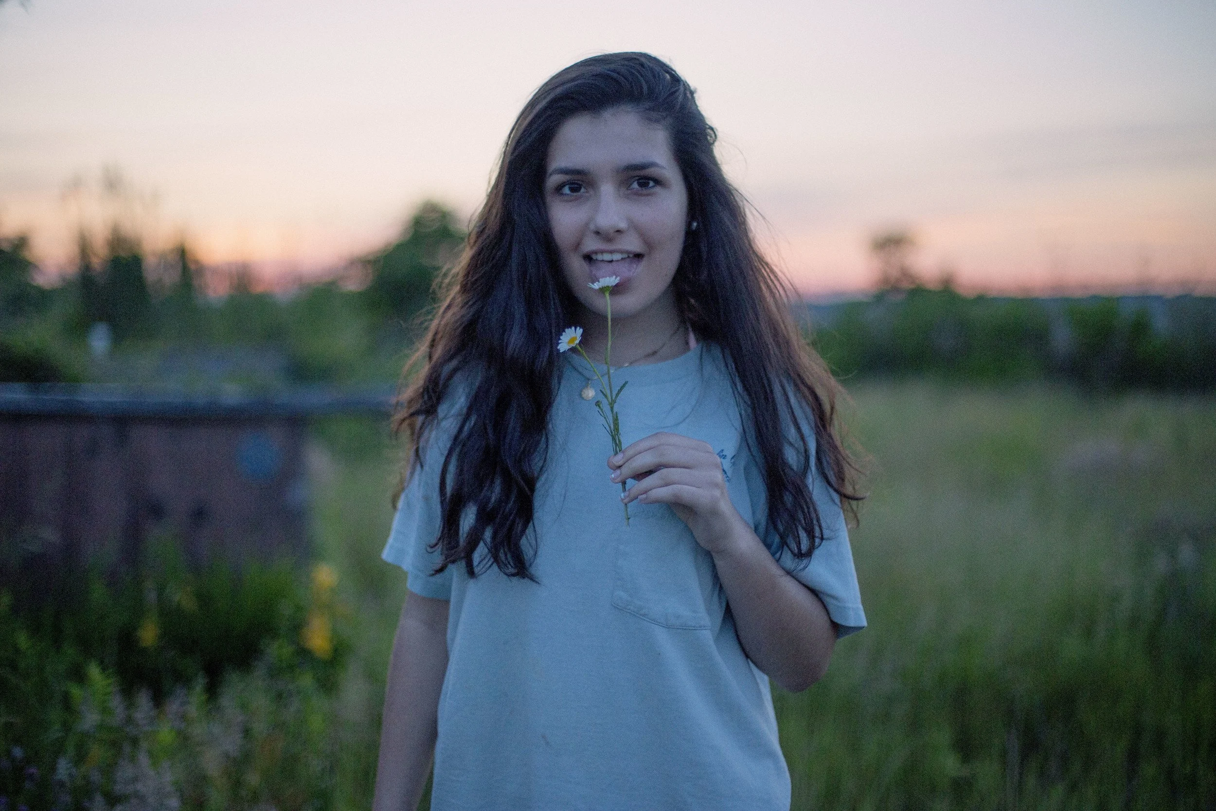 A young woman with long dark hair holding a small daisy flower to her lips outdoors at sunset, with a blurred green landscape in the background.