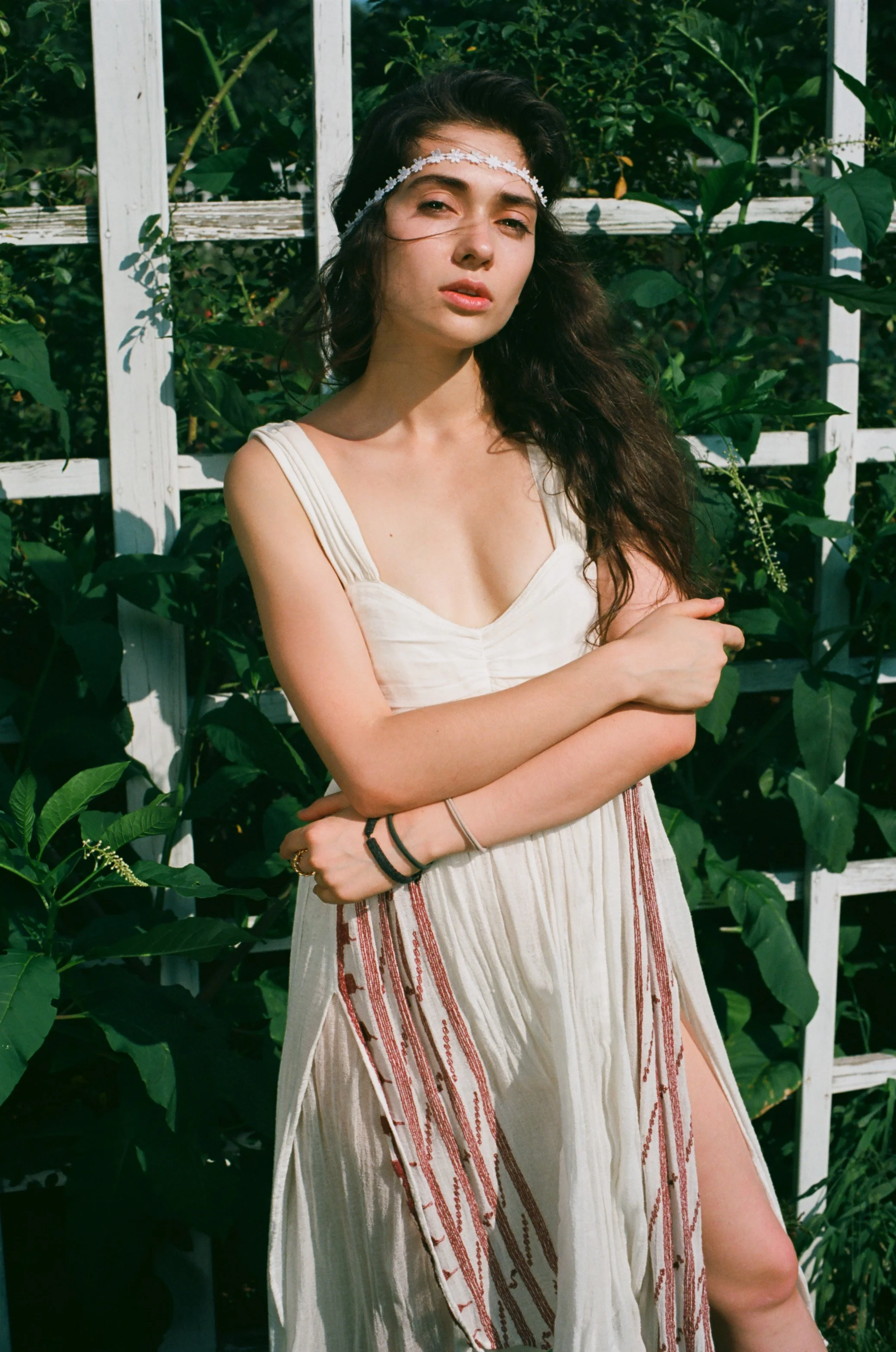 Young woman in a white dress with flower headband, standing outdoors with greenery and a white trellis in the background. Photo edited by Mary Perrino.