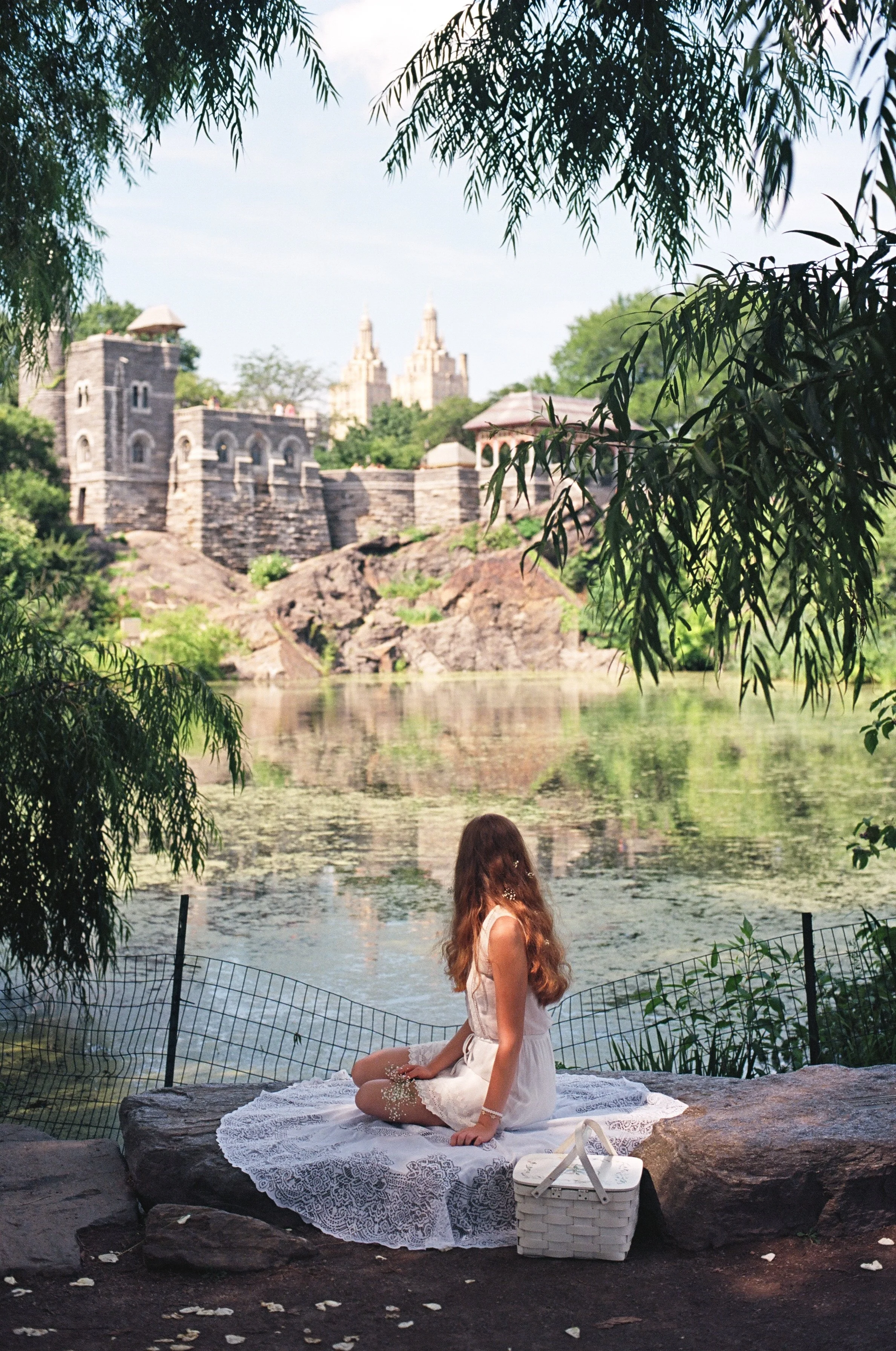 A woman with long wavy hair in a white dress sitting cross-legged on a lace blanket by a lake, surrounded by trees, with a castle-like building and towers in the background.