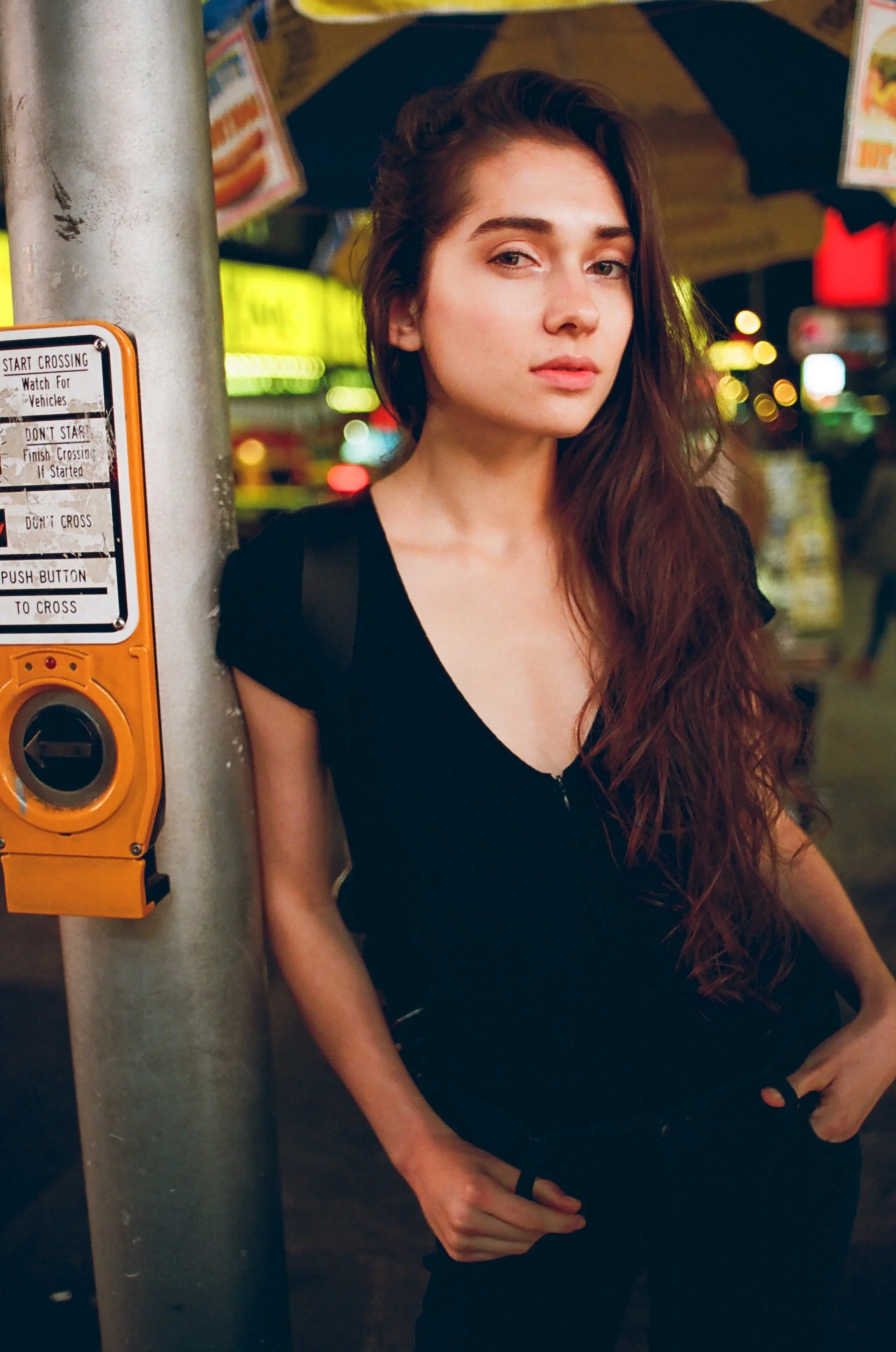 A young woman with long brown hair standing on a city street at night, next to a pedestrian crossing button, with neon lights and signs in the background.