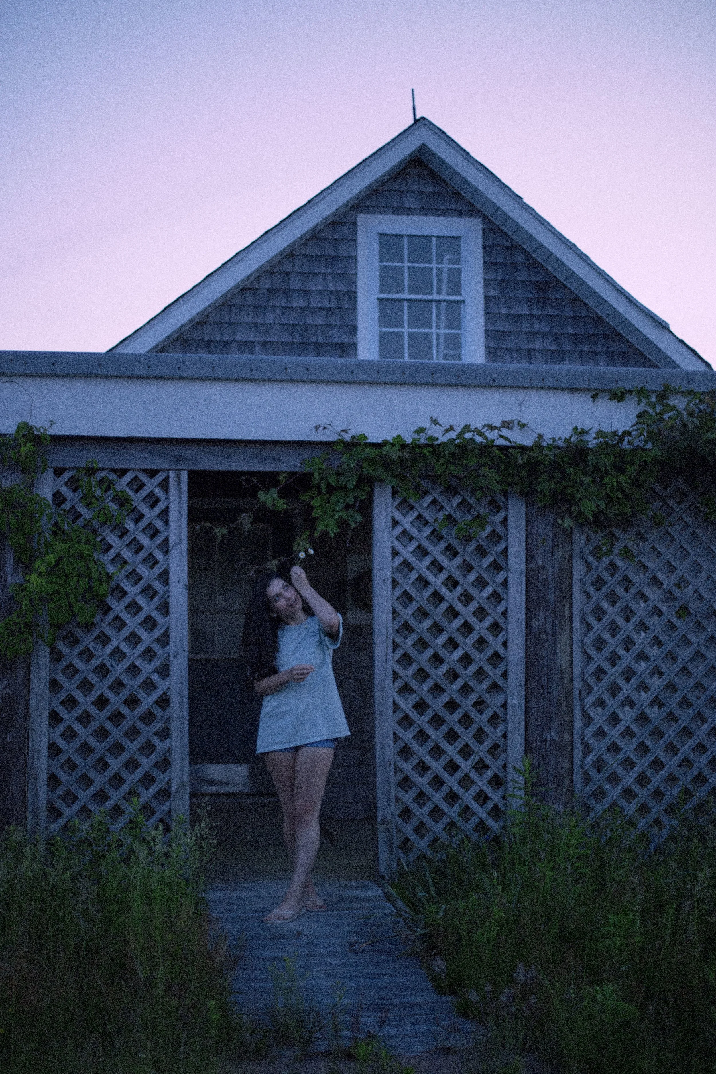 A woman standing at the entrance of a wooden porch under a covered area next to a house with a steep roof and a window, during twilight or early evening. Photo edited by Mary Perrino.