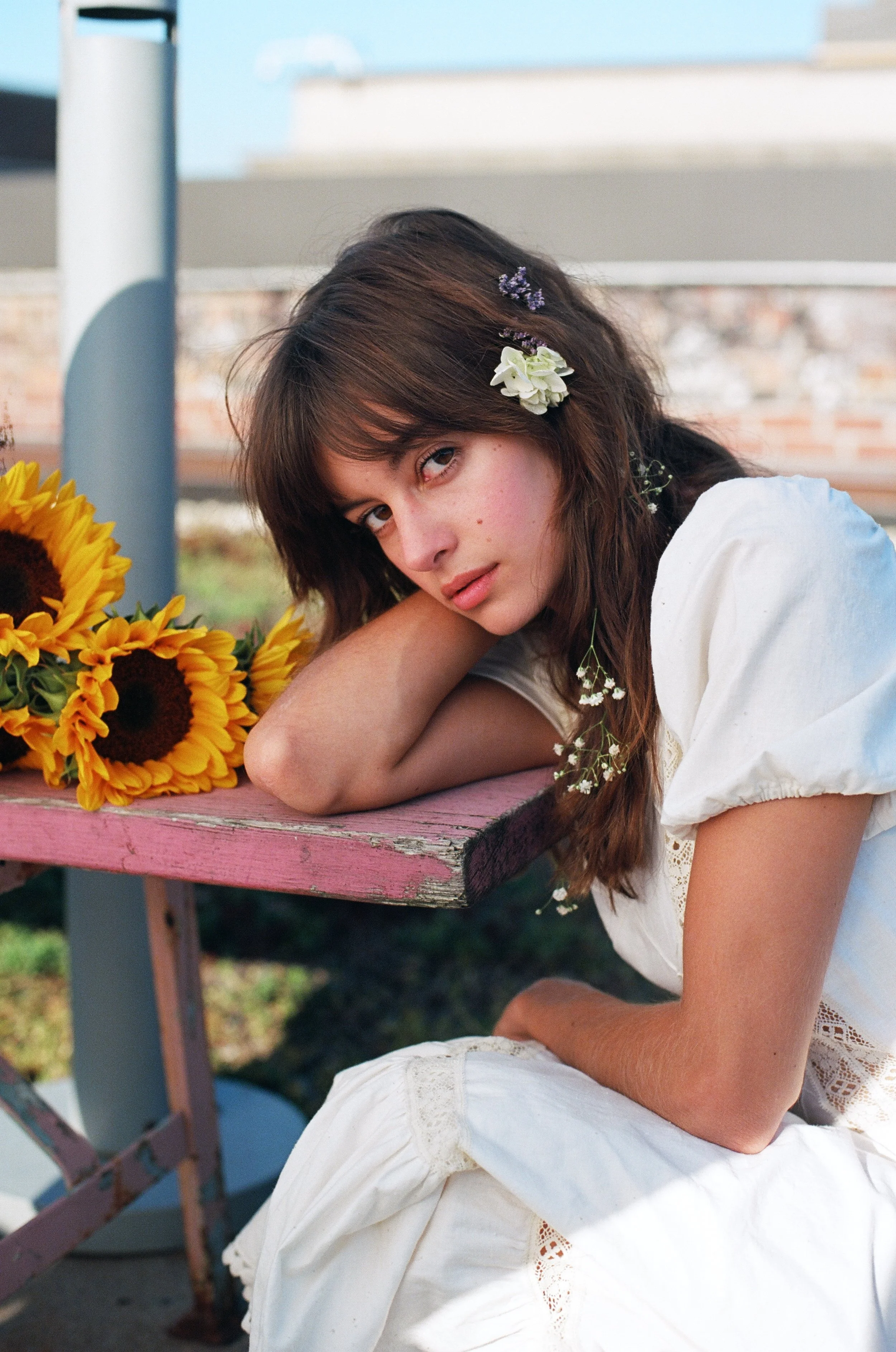 A young woman with long brown hair wearing a white dress, resting her head on her arm while sitting by a pink wooden table with sunflowers and small white flowers in her hair, outside on a sunny day.