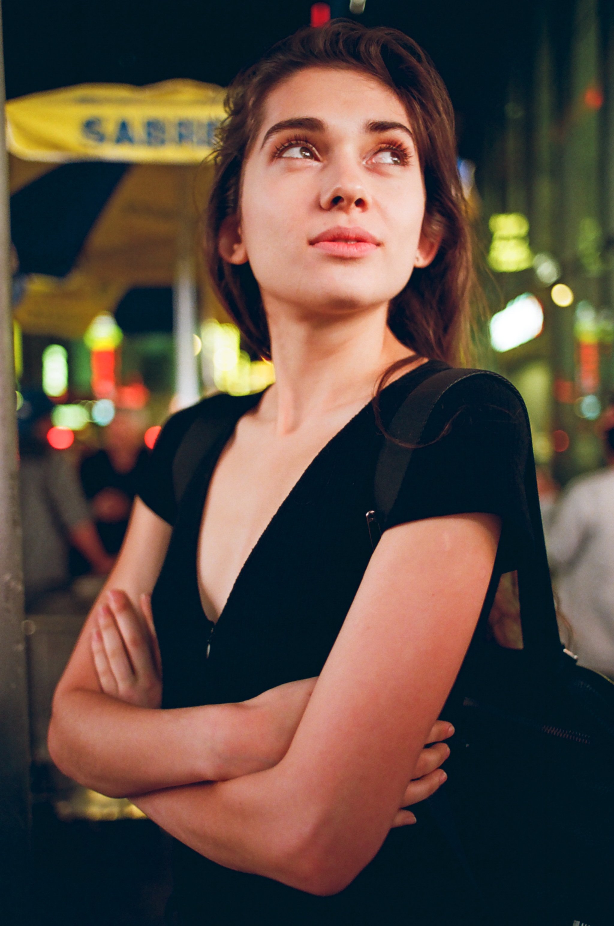 A young woman with shoulder-length brown hair, crossing her arms, standing outdoors at night with colorful neon signs and blurred city lights in the background.
