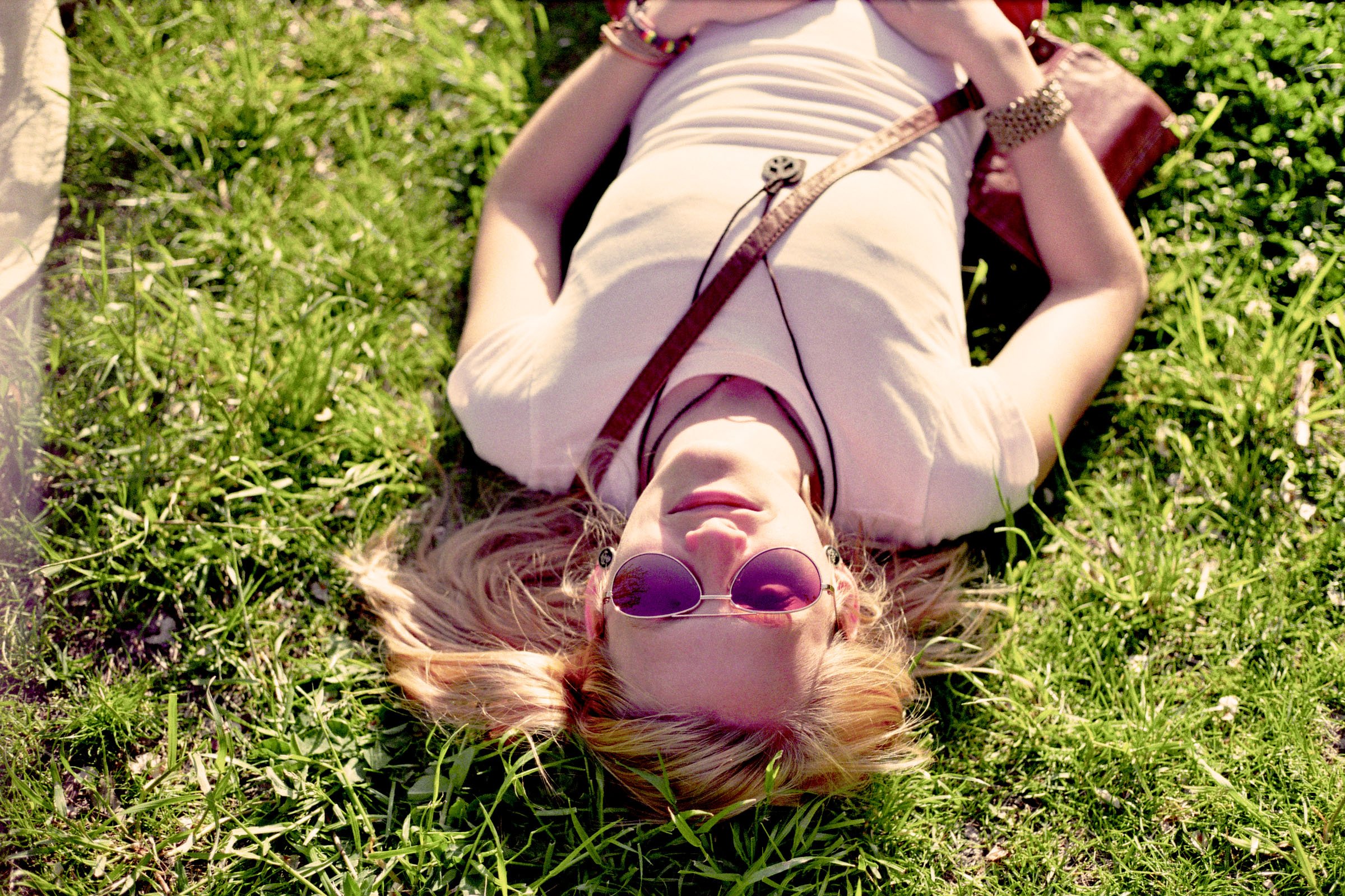 Woman lying on grass with sunglasses, wearing a light-colored dress, surrounded by green grass and small white flowers.