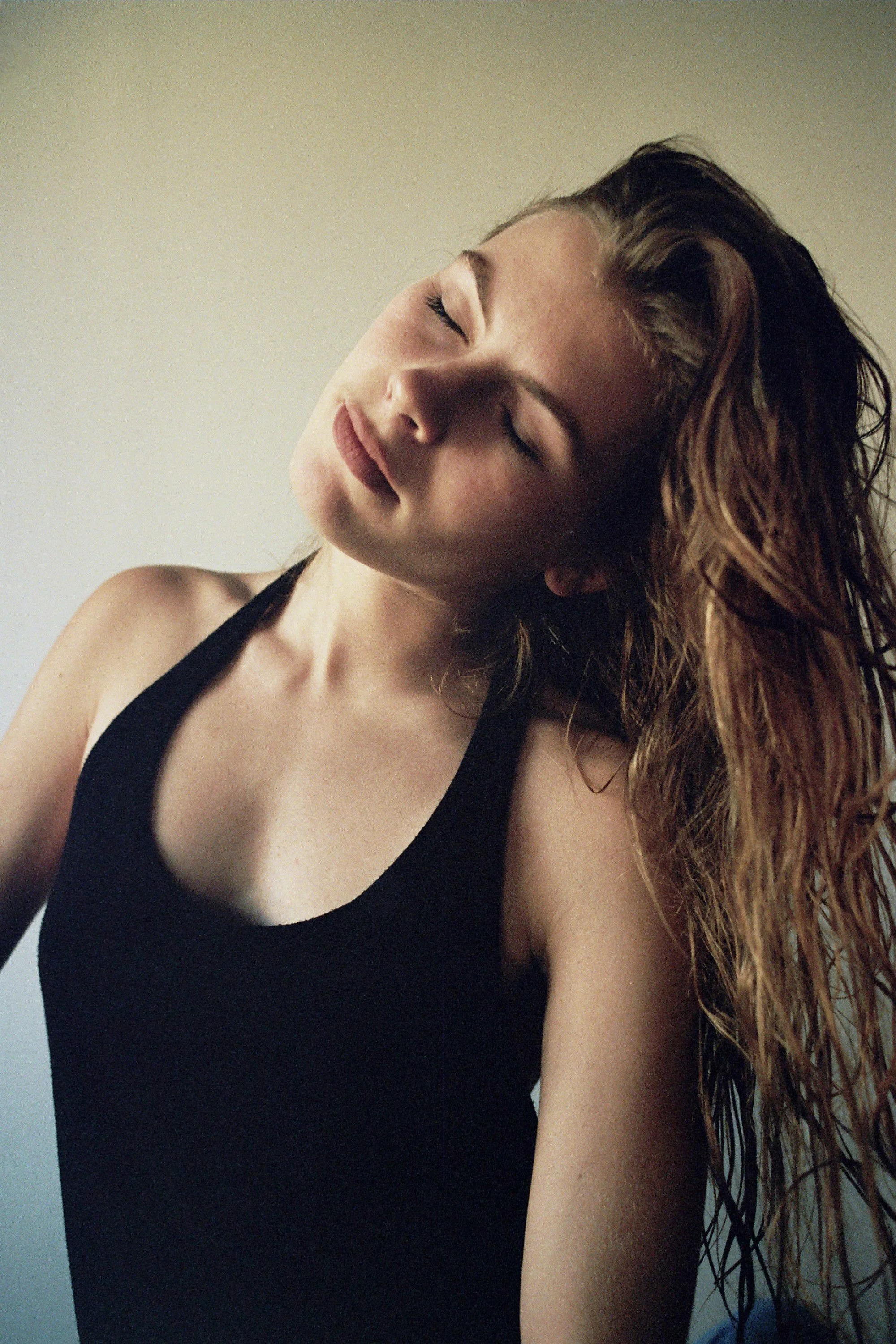 A young woman with long, wavy hair and closed eyes, wearing a black tank top, tilting her head with a peaceful expression.