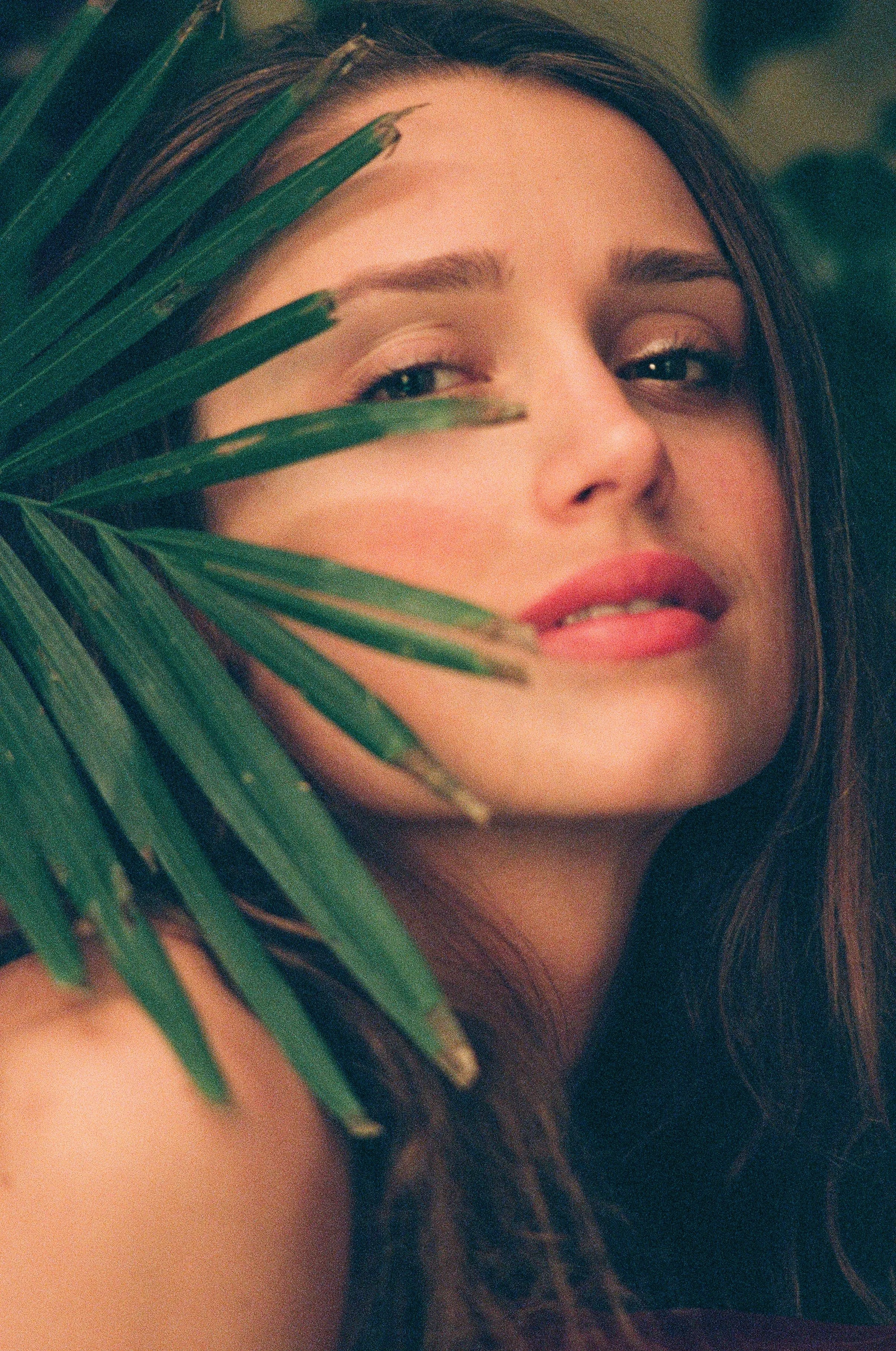Close-up portrait of a woman with long brown hair, partially hidden behind green palm leaves, with a soft, natural expression. Photo edited by Mary Perrino.