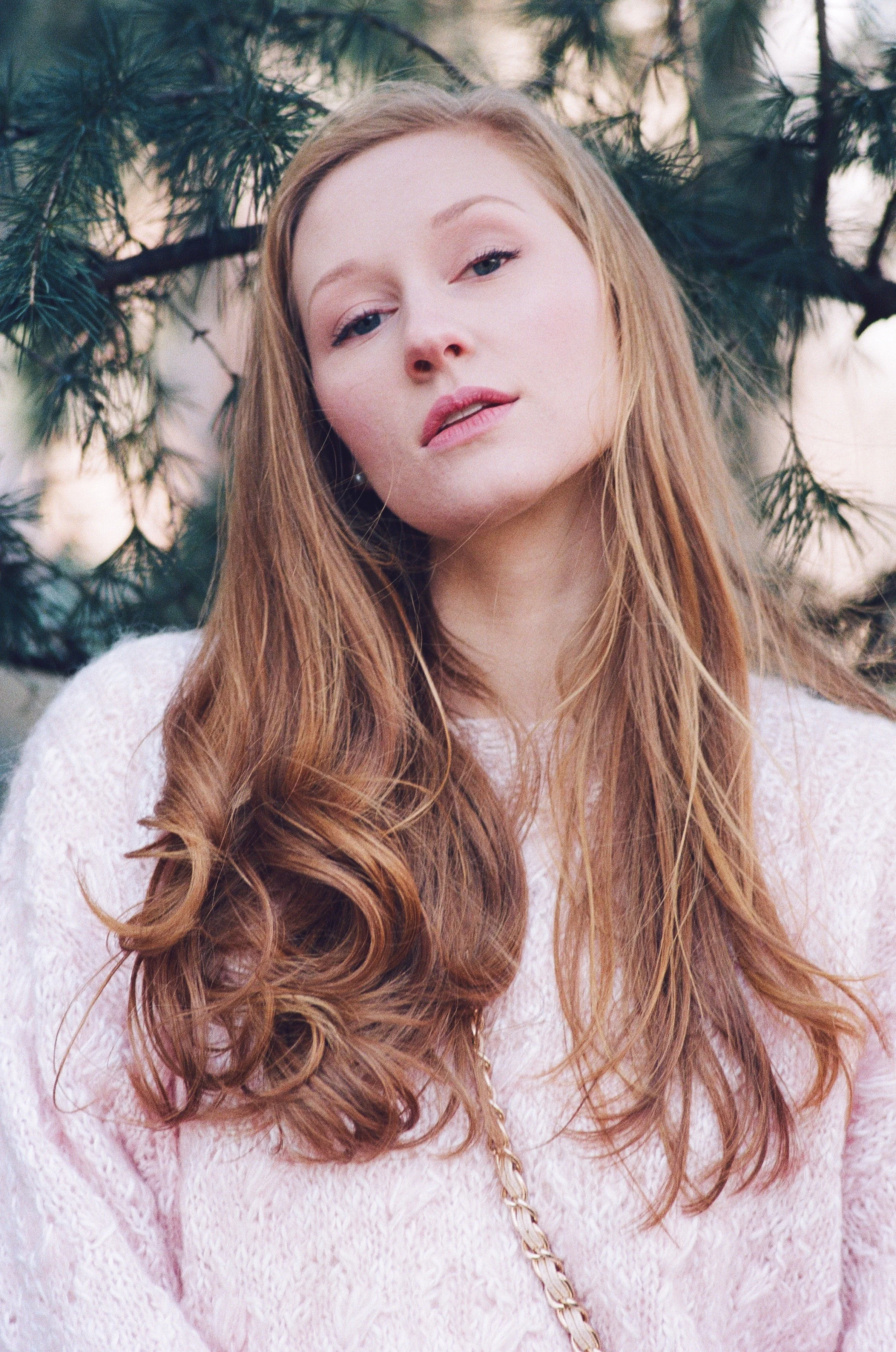 A woman with long red hair wearing a pink sweater stands outdoors, with pine tree branches in the background.