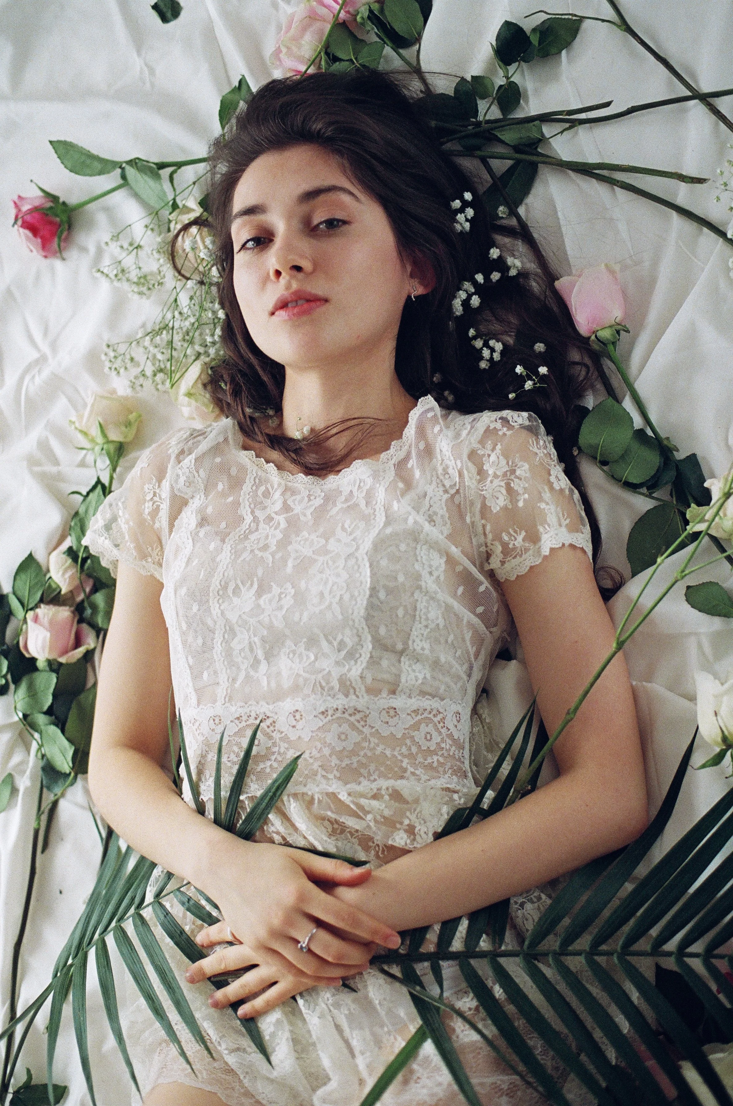 Young woman lying on a white sheet, surrounded by pink and white roses and green leaves, wearing a lace dress.