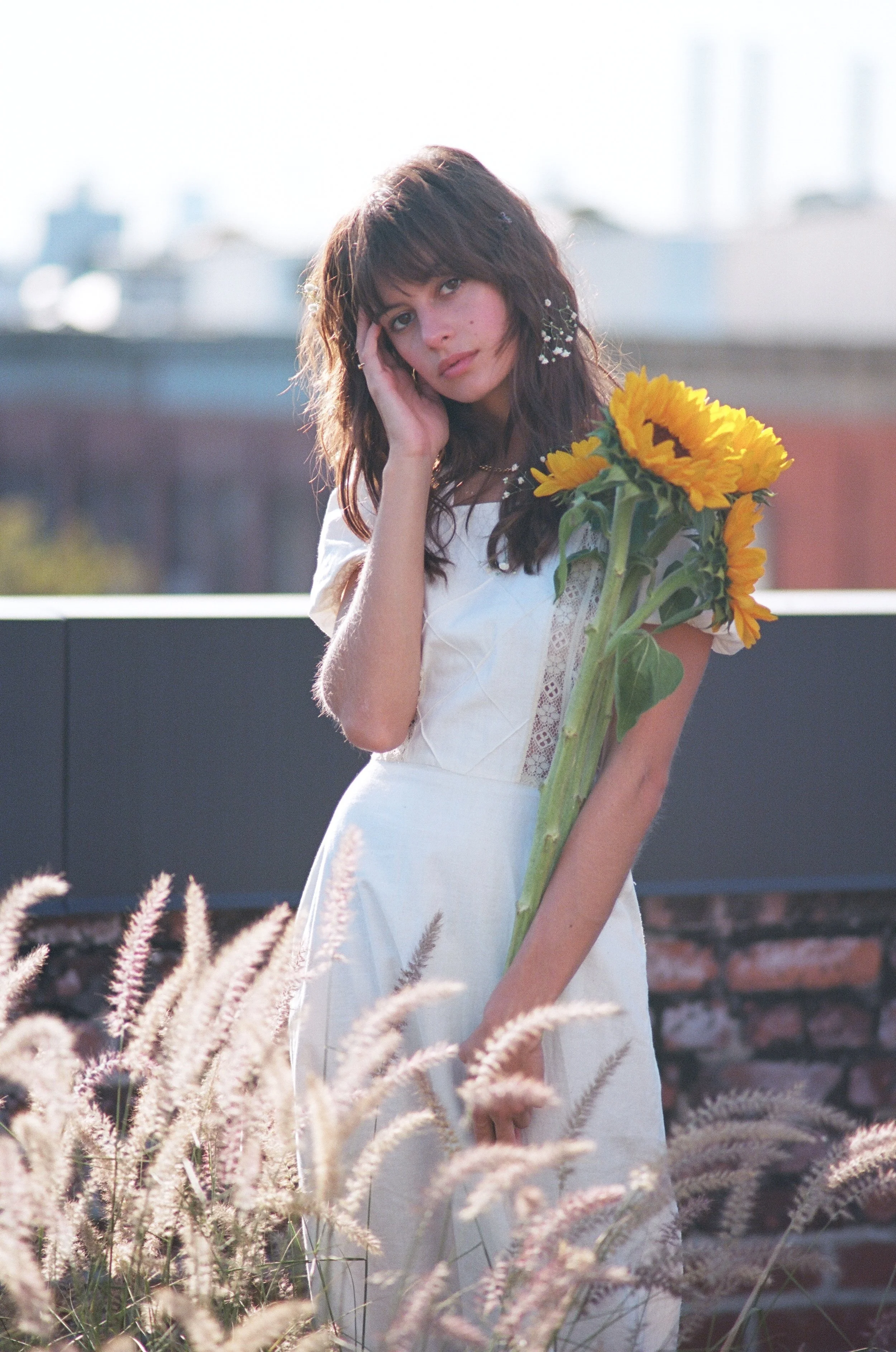 A young woman stands outdoors holding a bunch of sunflowers, dressed in a white dress with lace details, with a cityscape and plants in the background.