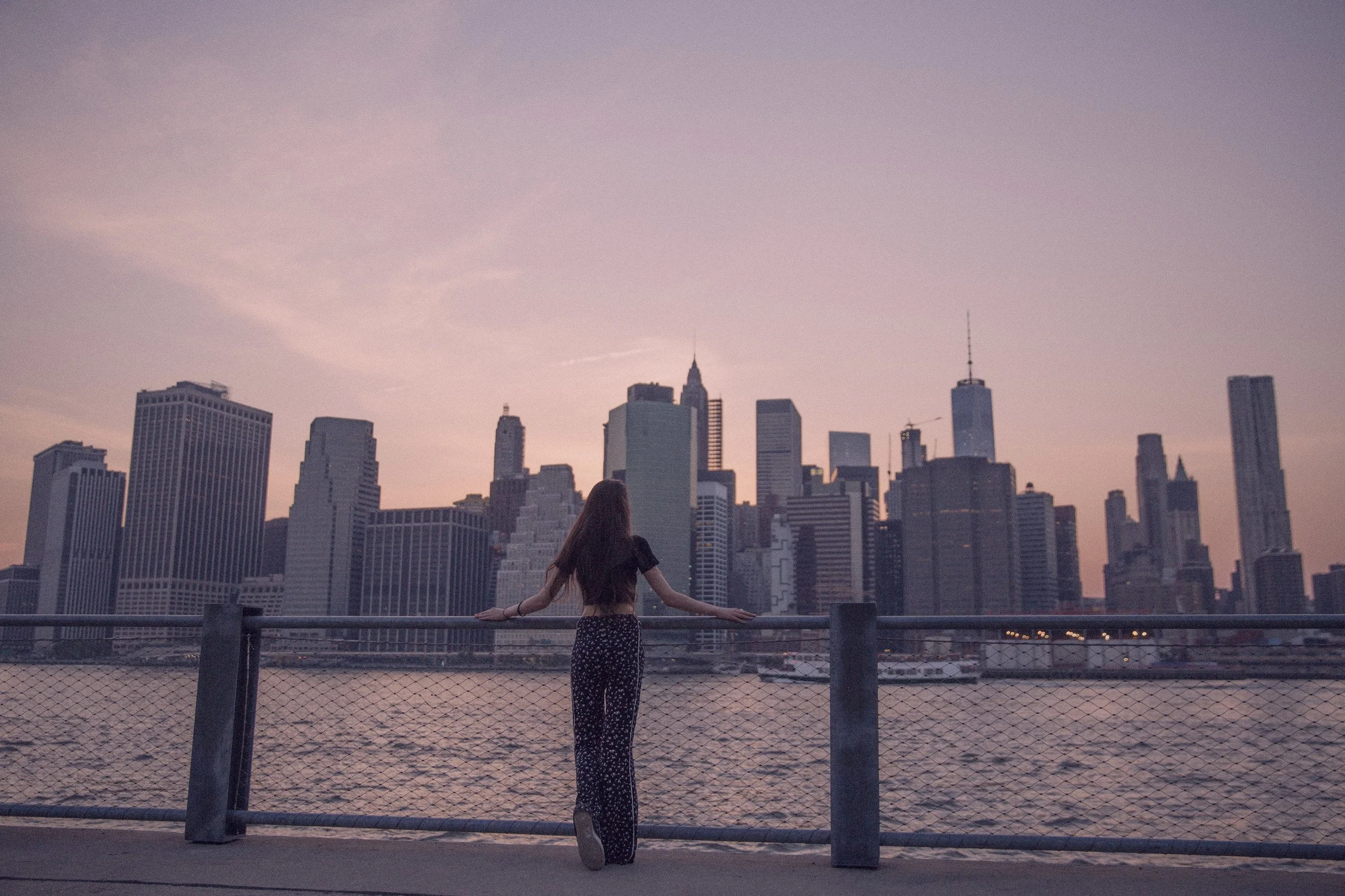 A woman standing on a waterfront promenade, facing a city skyline during sunset.