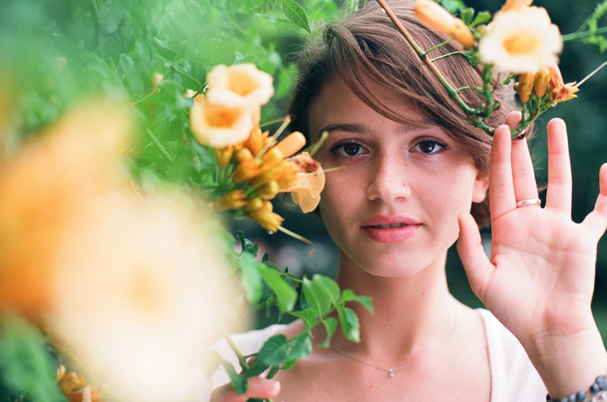 A woman with short brown hair and light makeup among orange and yellow flowers, raising her hand near her face with a natural background.