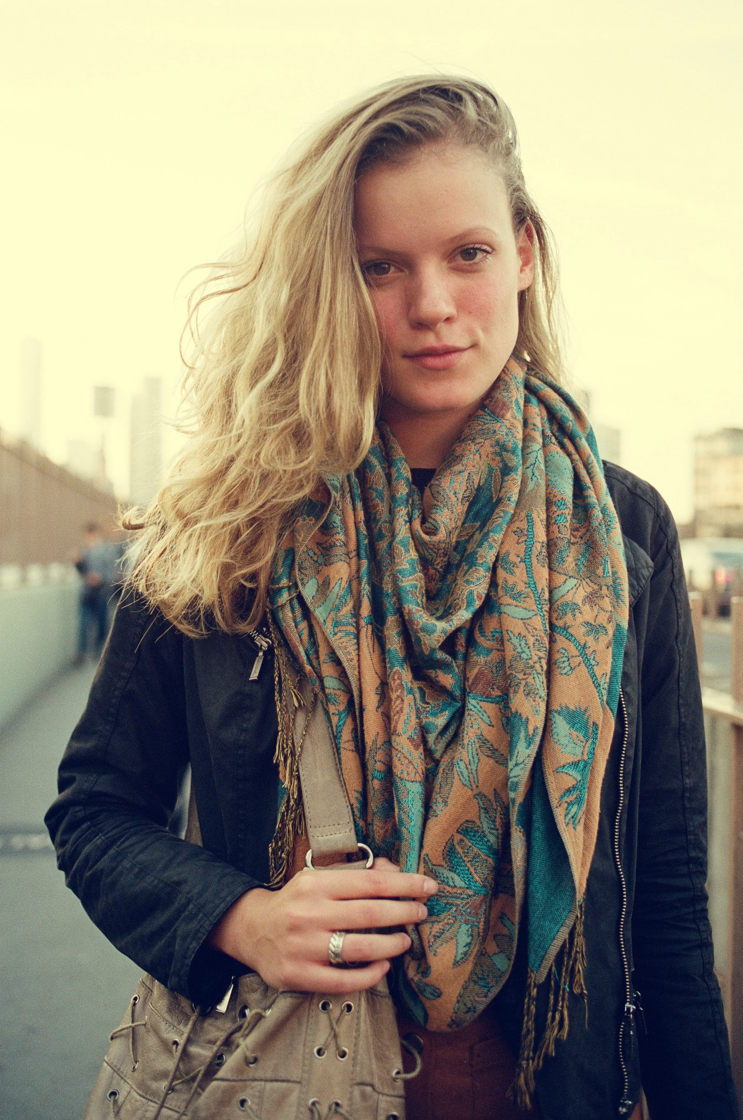 A young woman with long blonde wavy hair standing outdoors, wearing a black leather jacket and a multicolored scarf, holding a beige purse, with a city skyline in the background.