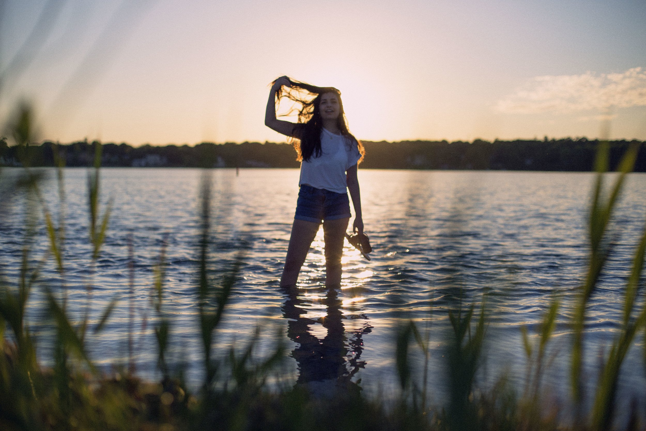 A woman standing in a lake at sunset, holding a hat, with tall grasses in the foreground and trees in the background.