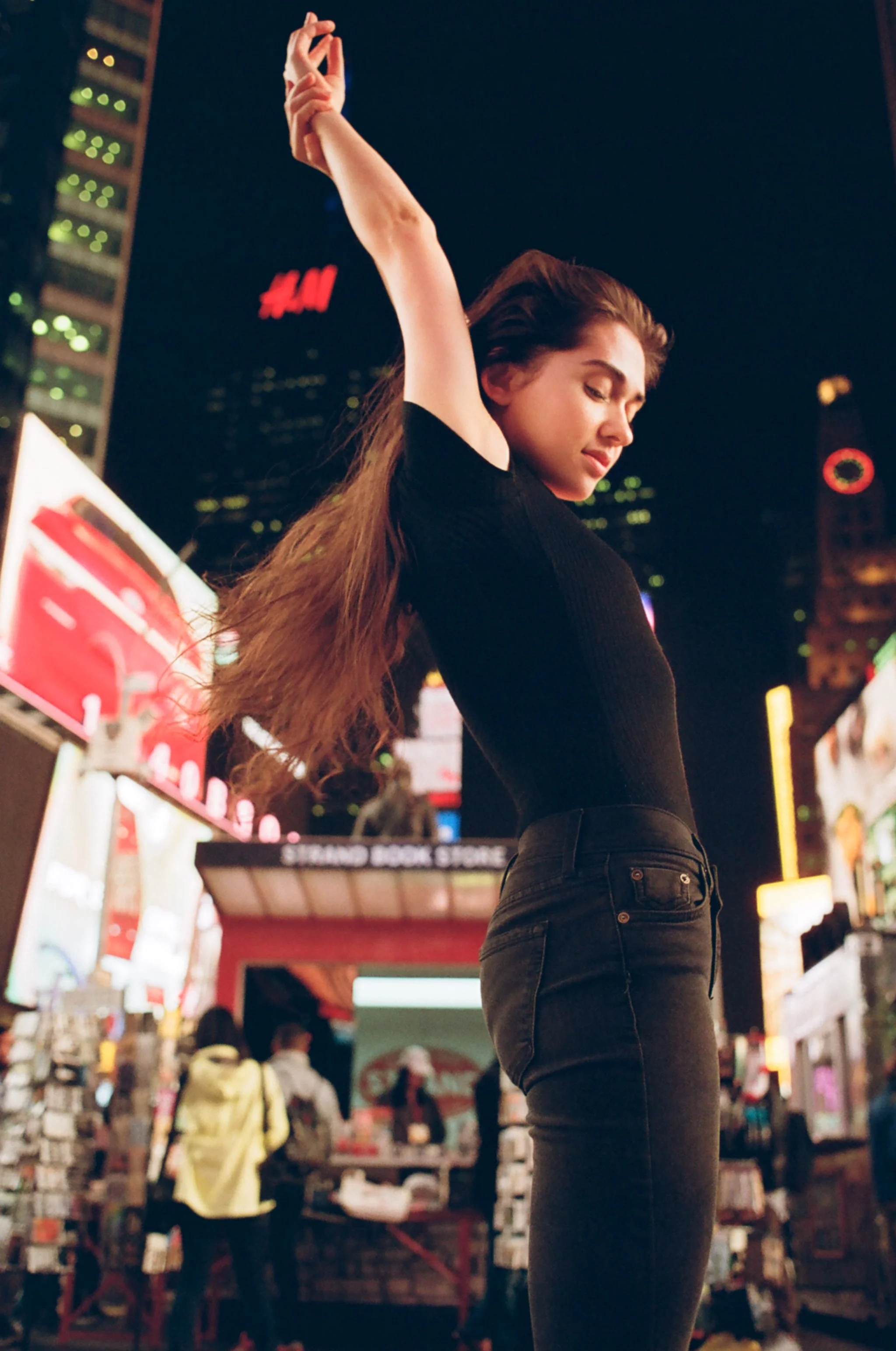 A woman with long hair in a black top and black jeans stretching her arms at night in Times Square, New York City, surrounded by bright billboards and digital advertisements.