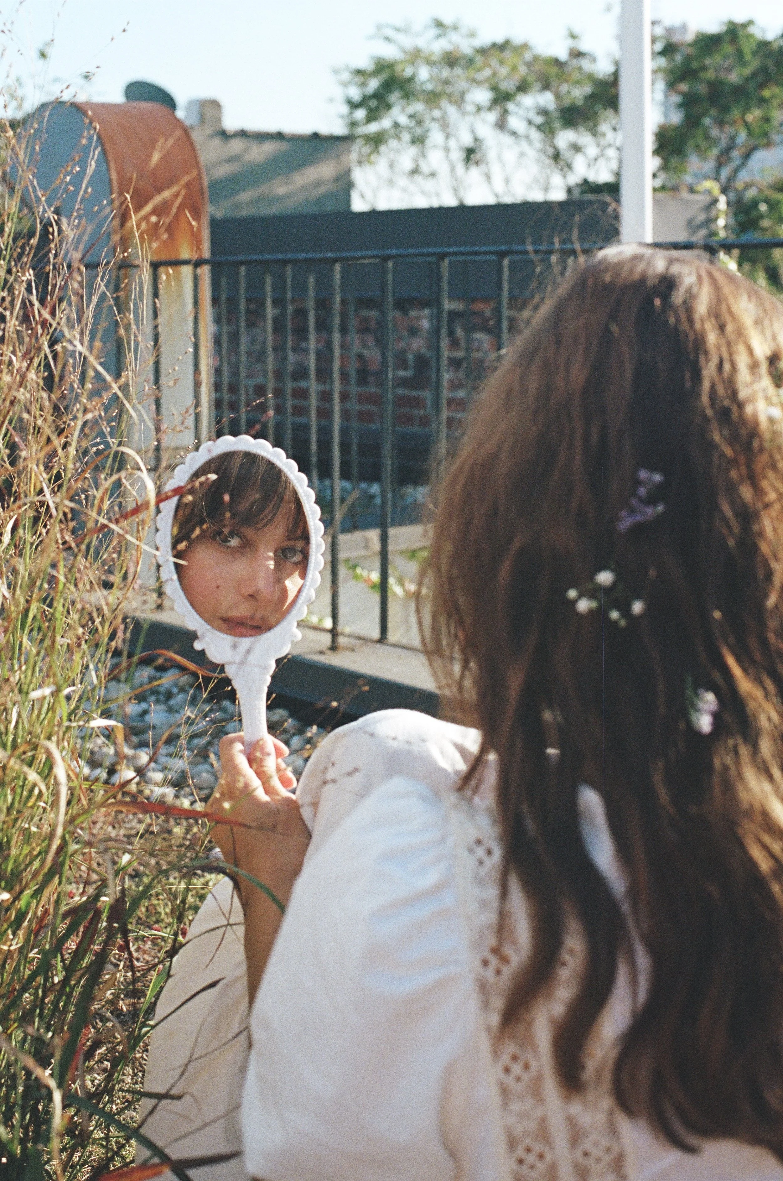 A young woman with curly brown hair, decorated with small purple and white flowers, is sitting outdoors, holding a white hand mirror with a scalloped edge, reflecting her face.