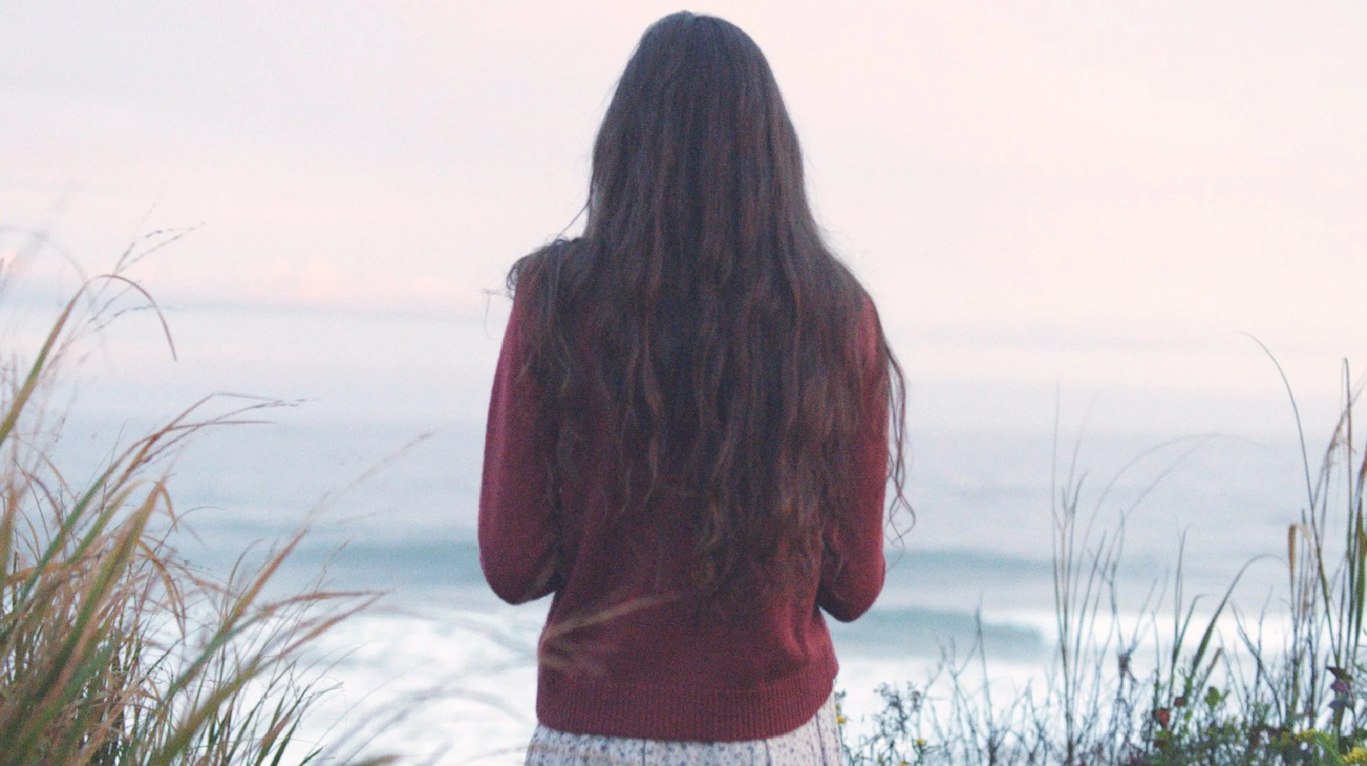 A woman with long dark hair standing on a grassy shoreline facing the ocean, with waves and a cloudy sky in the background. Photo edited by Mary Perrino.