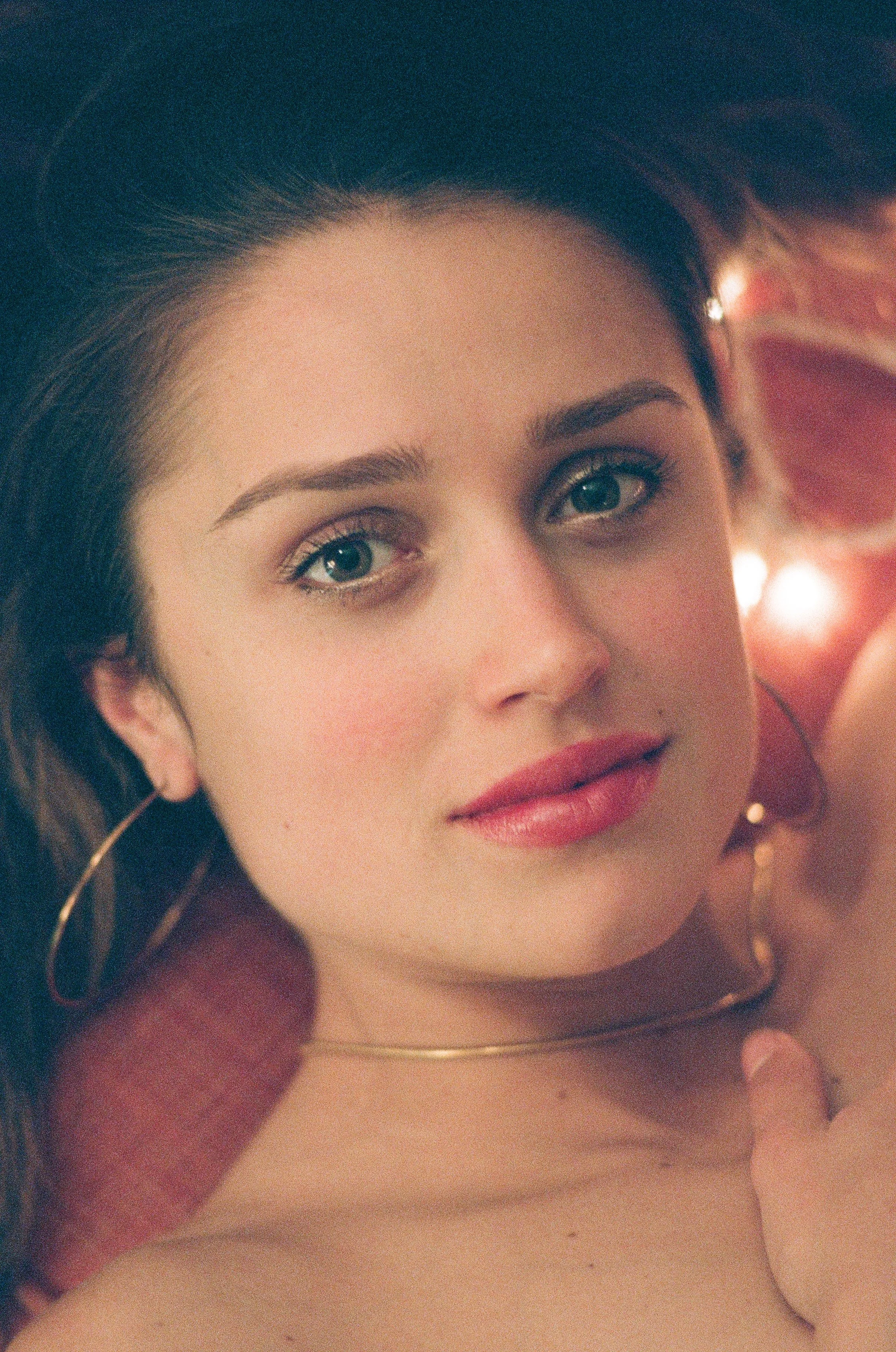 Close-up of a woman with brown hair, blue eyes, pink lipstick, gold hoop earrings, and a gold necklace, lying on a red pillow with soft lighting.