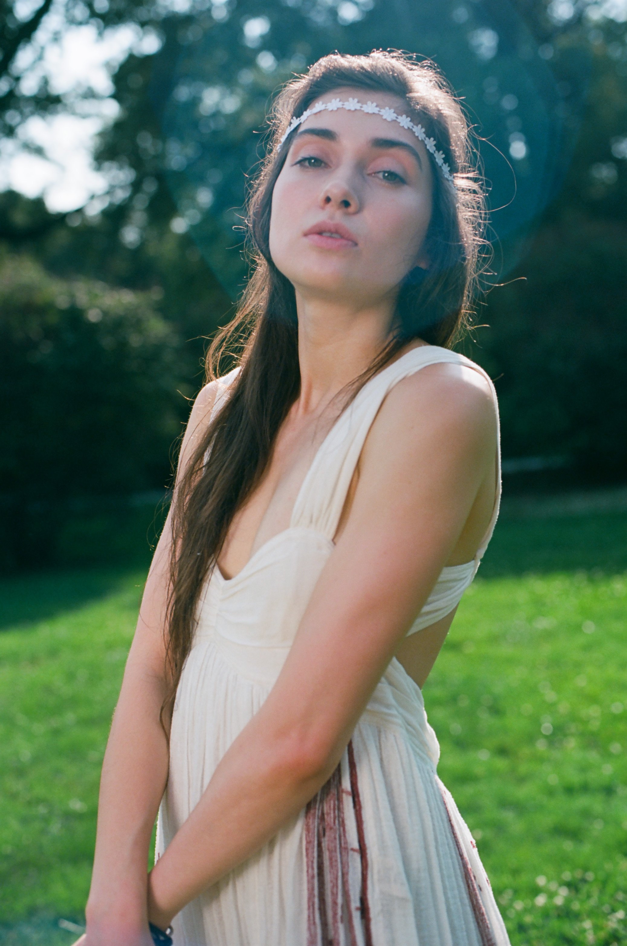 Young woman with long brown hair wearing a white dress and a floral headband, standing outdoors with green grass and trees in the background.