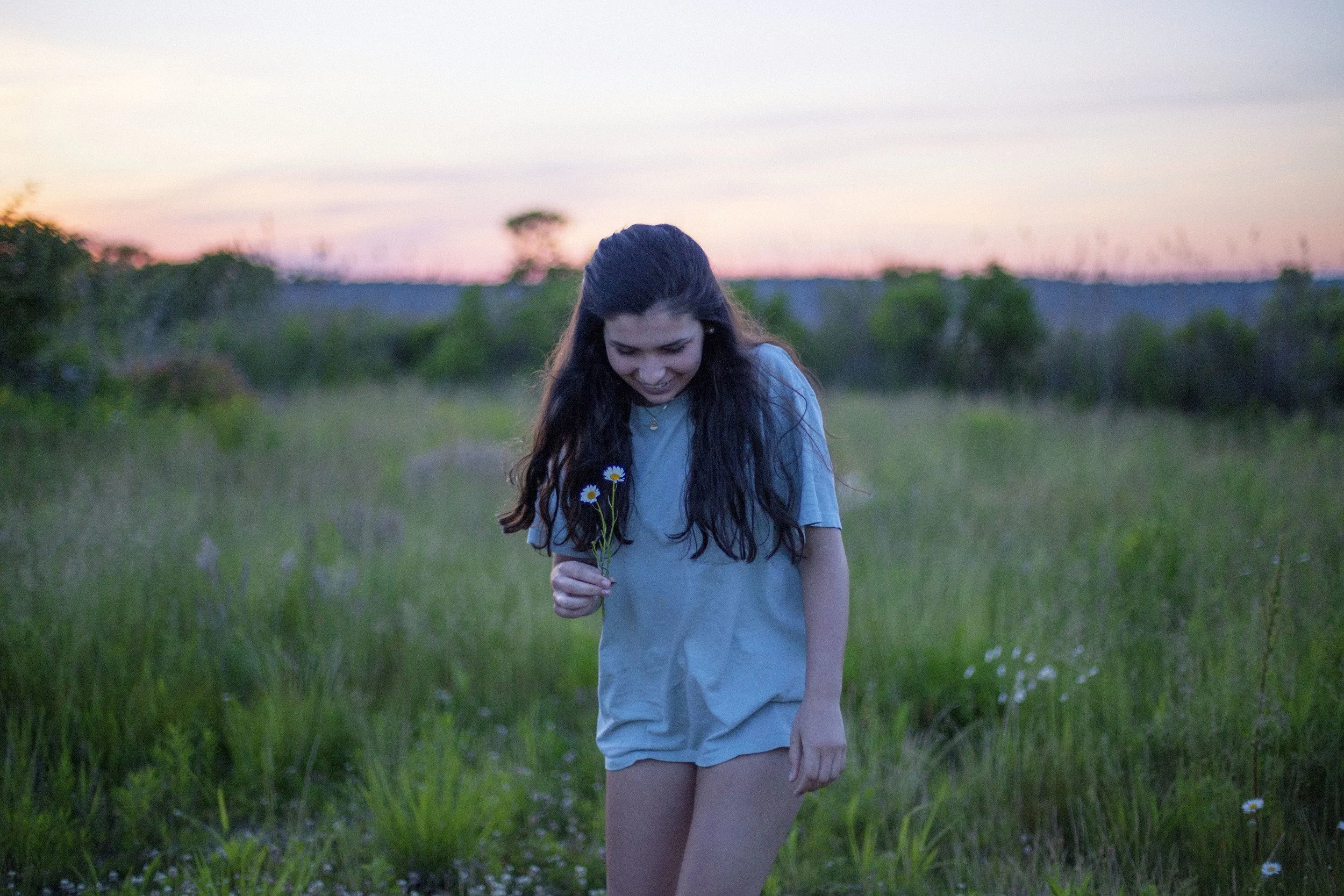 A young woman with long dark hair is standing in a grassy field during sunset, holding a small bouquet of daisies and smiling.