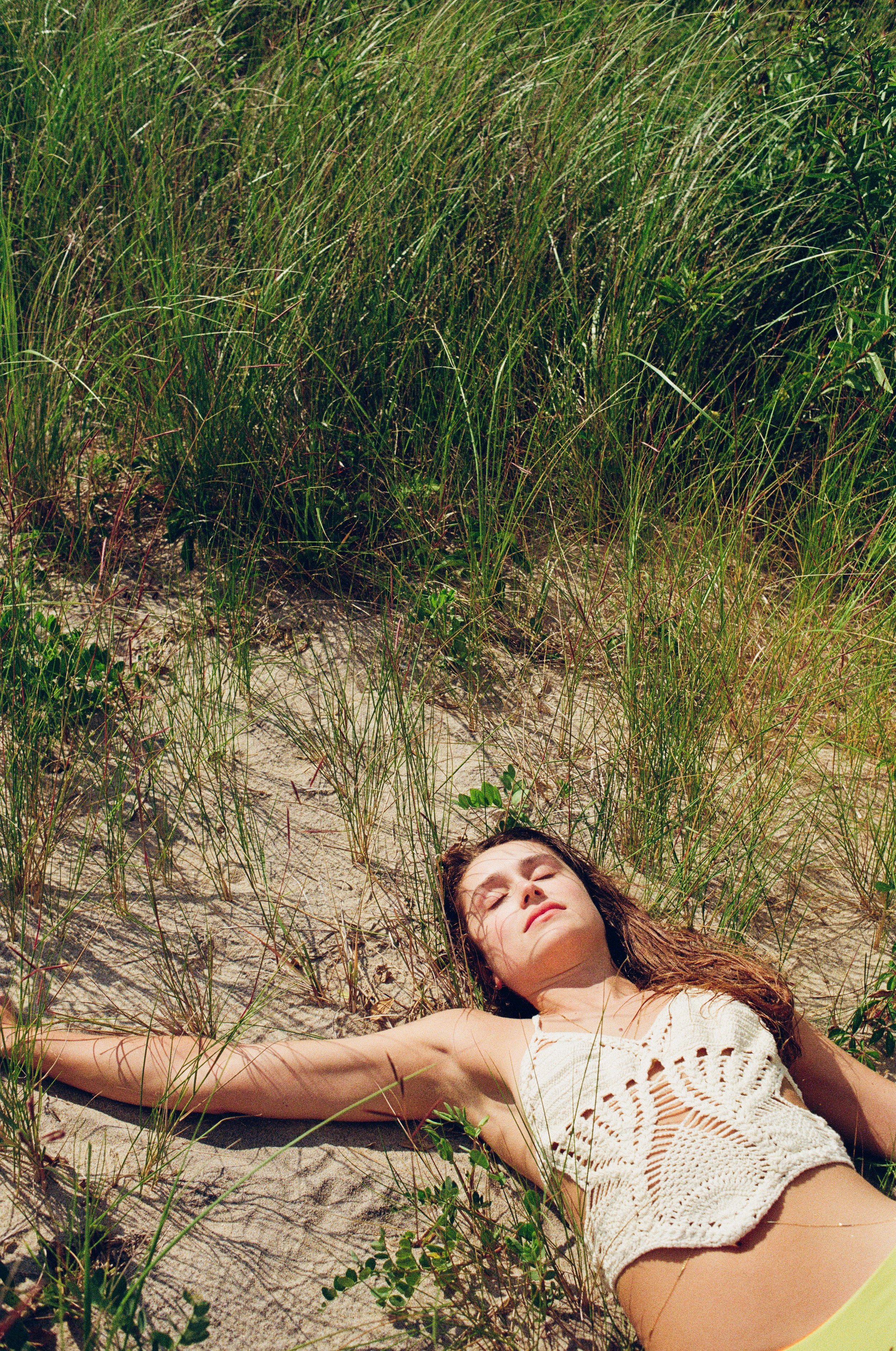 A woman relaxing on the sand at the beach, surrounded by tall grass, with eyes closed and wearing a sleeveless knitted top. Photo edited by Mary Perrino.