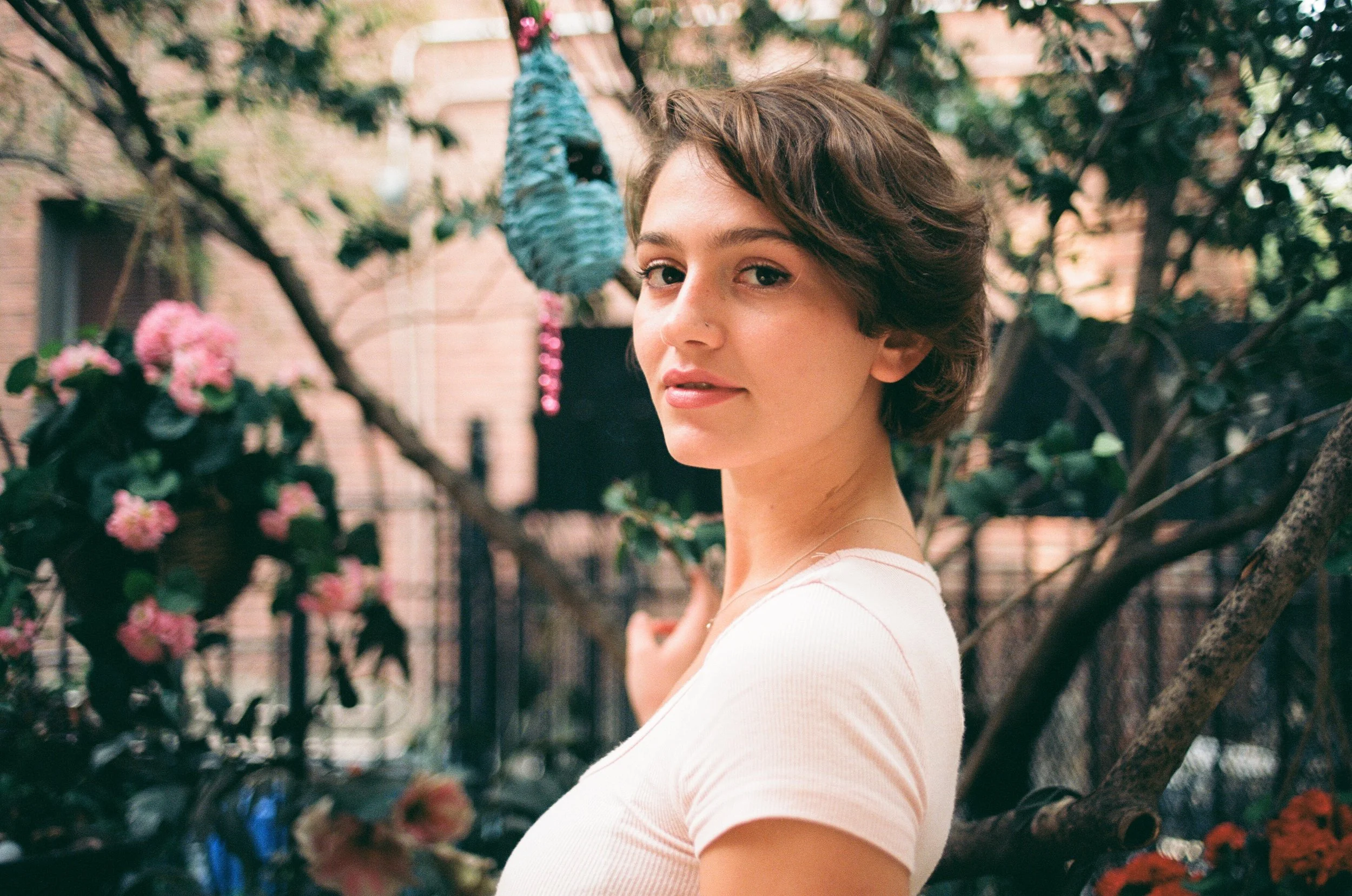 A young woman with short brown hair and a light pink shirt standing outdoors near a tree with pink and red flowers.