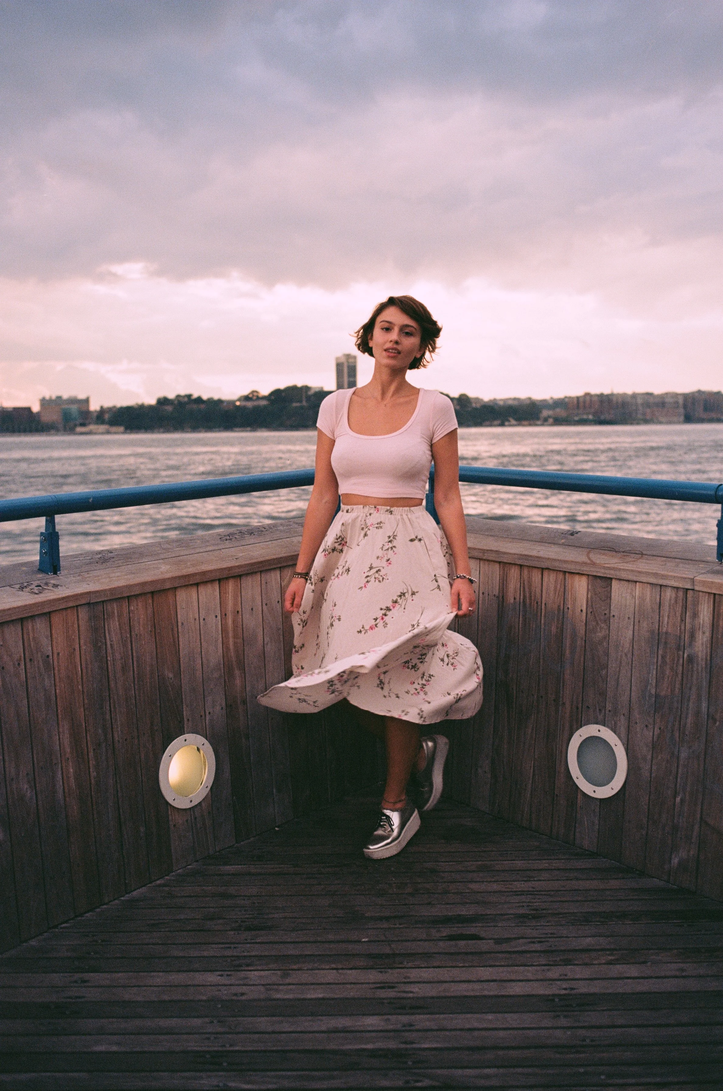 A young woman standing on a wooden dock by the water, with city skyline and cloudy sky in the background, wearing a white crop top, floral skirt, and sneakers.