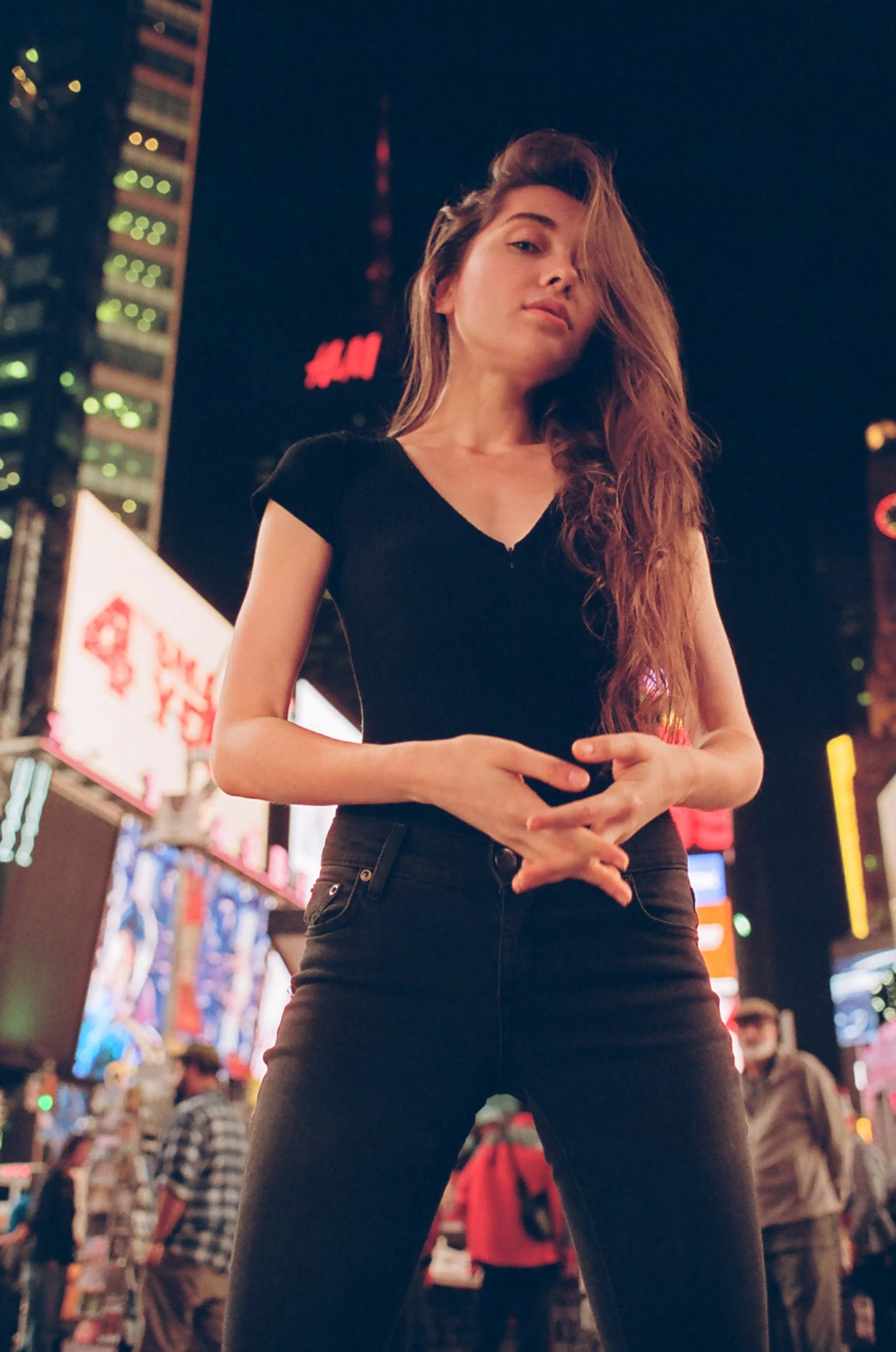 A young woman standing in Times Square at night, surrounded by bright billboards and city lights. She has long hair, wears a black top and pants, and looks confidently at the camera.