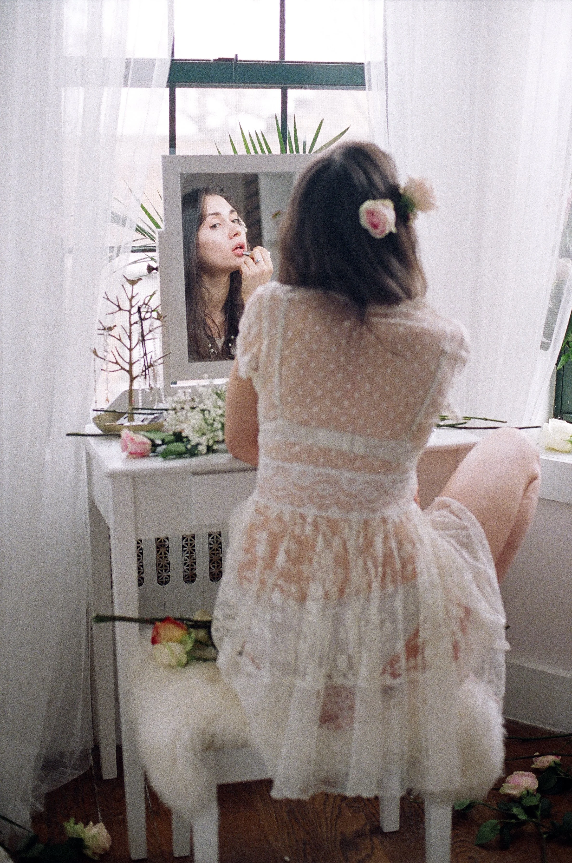 A woman in a lace dress with a floral hair accessory sitting in front of a mirror applying lipstick, with makeup and flowers on a white vanity.