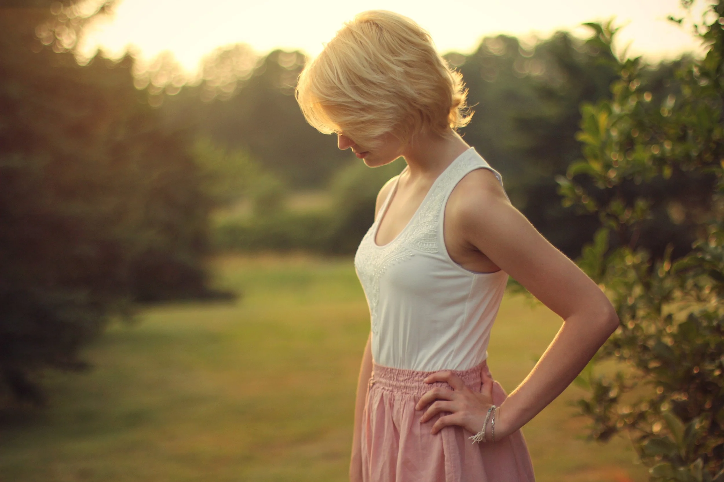 A woman with blonde hair wearing a white sleeveless top and pink skirt standing outdoors in a garden or park during sunset.