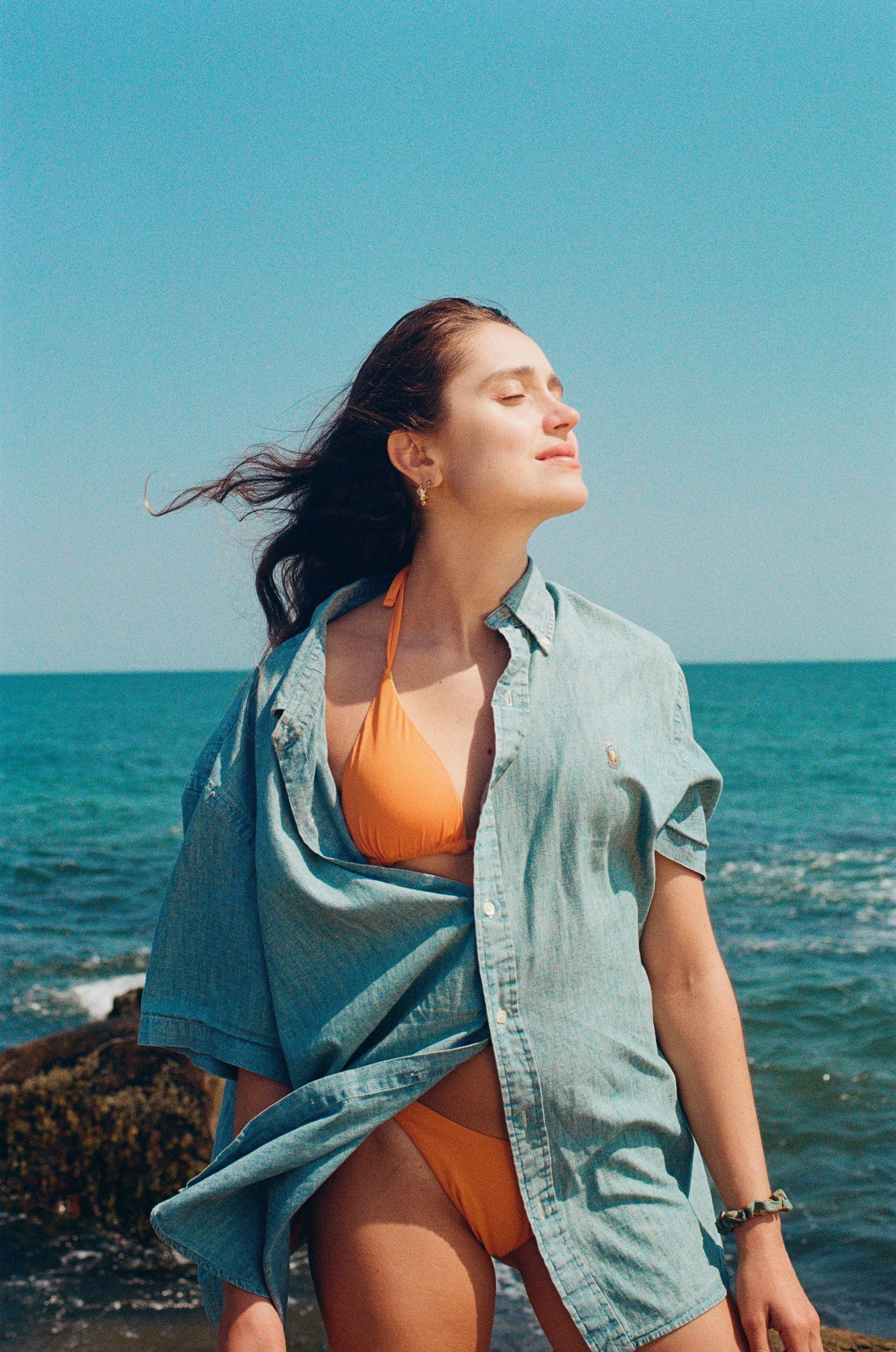 Woman wearing an orange bikini and a denim shirt standing on a beach with the ocean in the background, enjoying sunny weather. Photo edited by Mary Perrino.