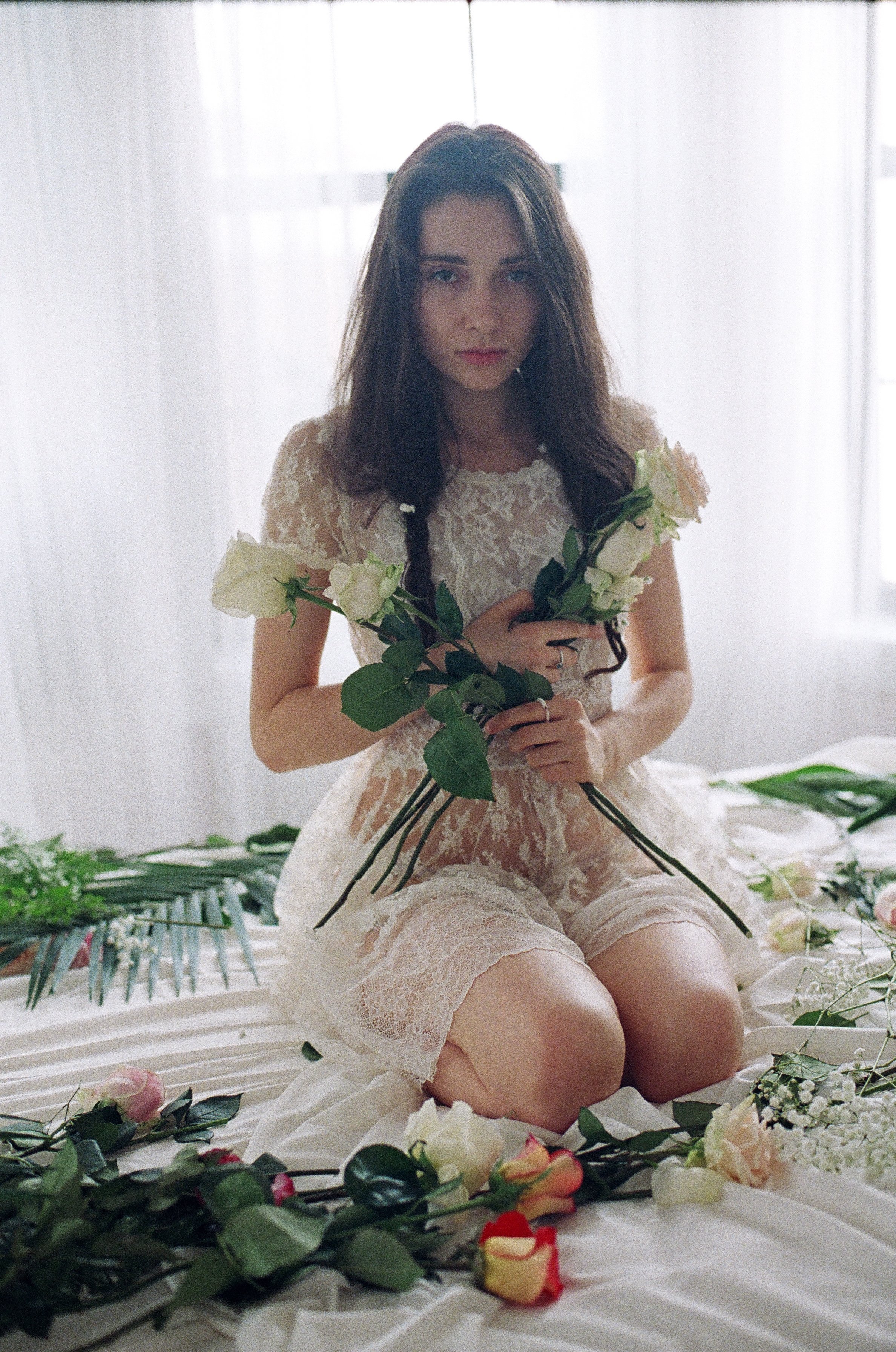 A young woman with long dark hair sitting on a bed covered with white fabric and decorated with roses, holding a bunch of white roses, in a softly lit room.