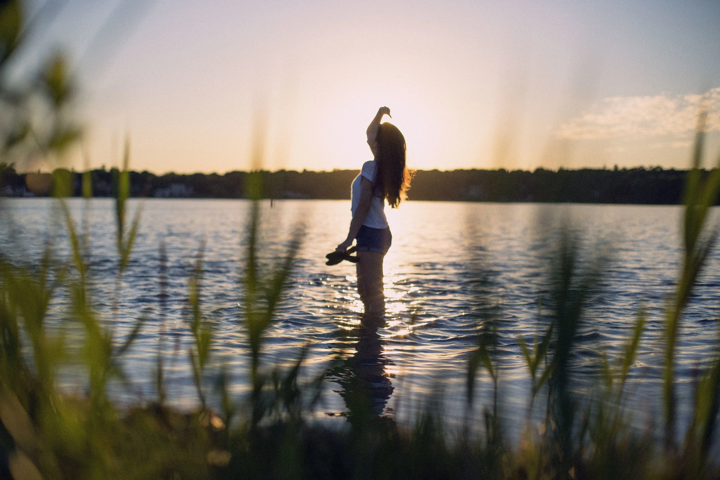 A woman holding a phone standing in water at sunset, with grass in the foreground and a distant tree line.