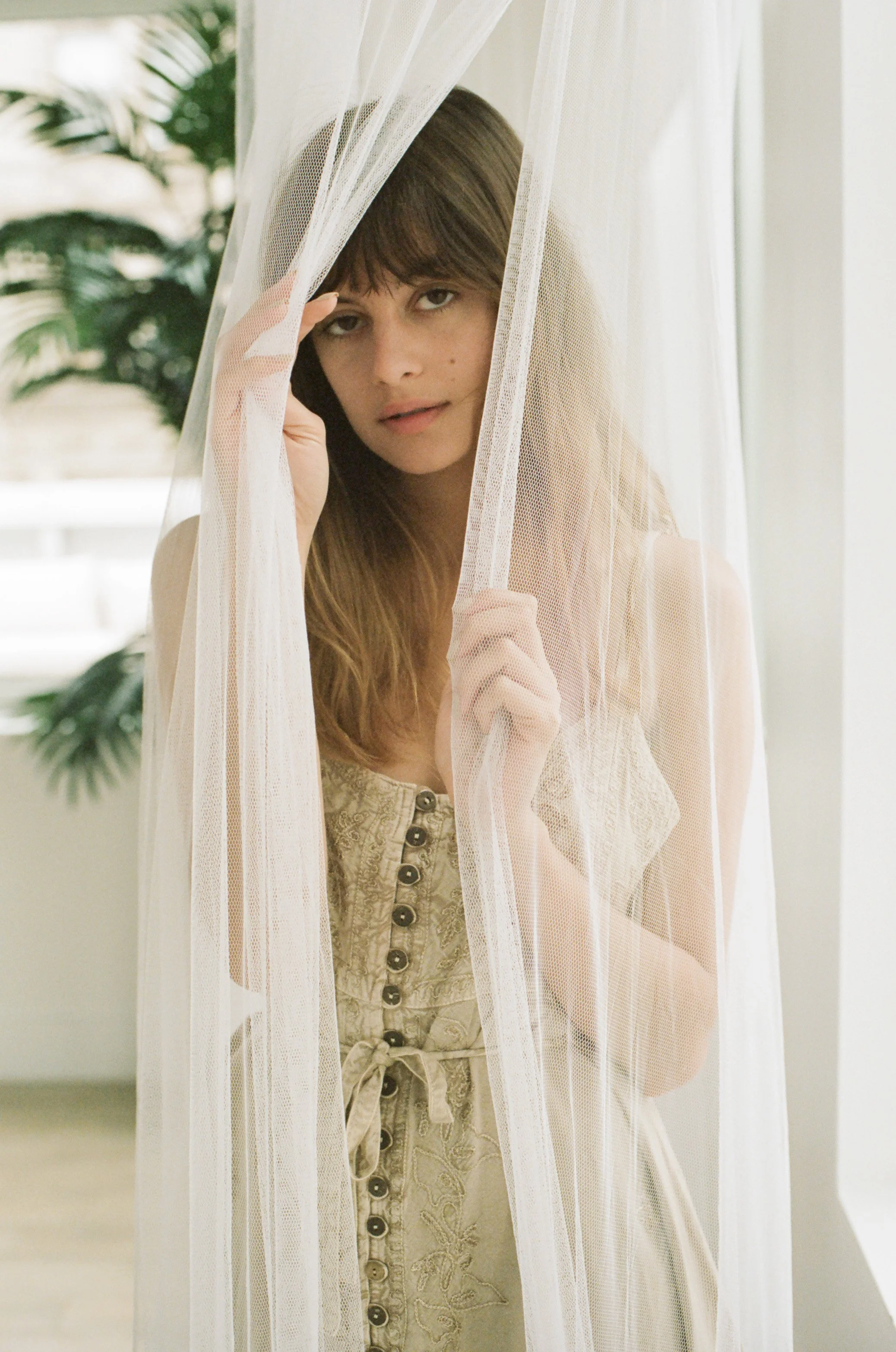 Young woman with long brown hair peeking through sheer white curtain.