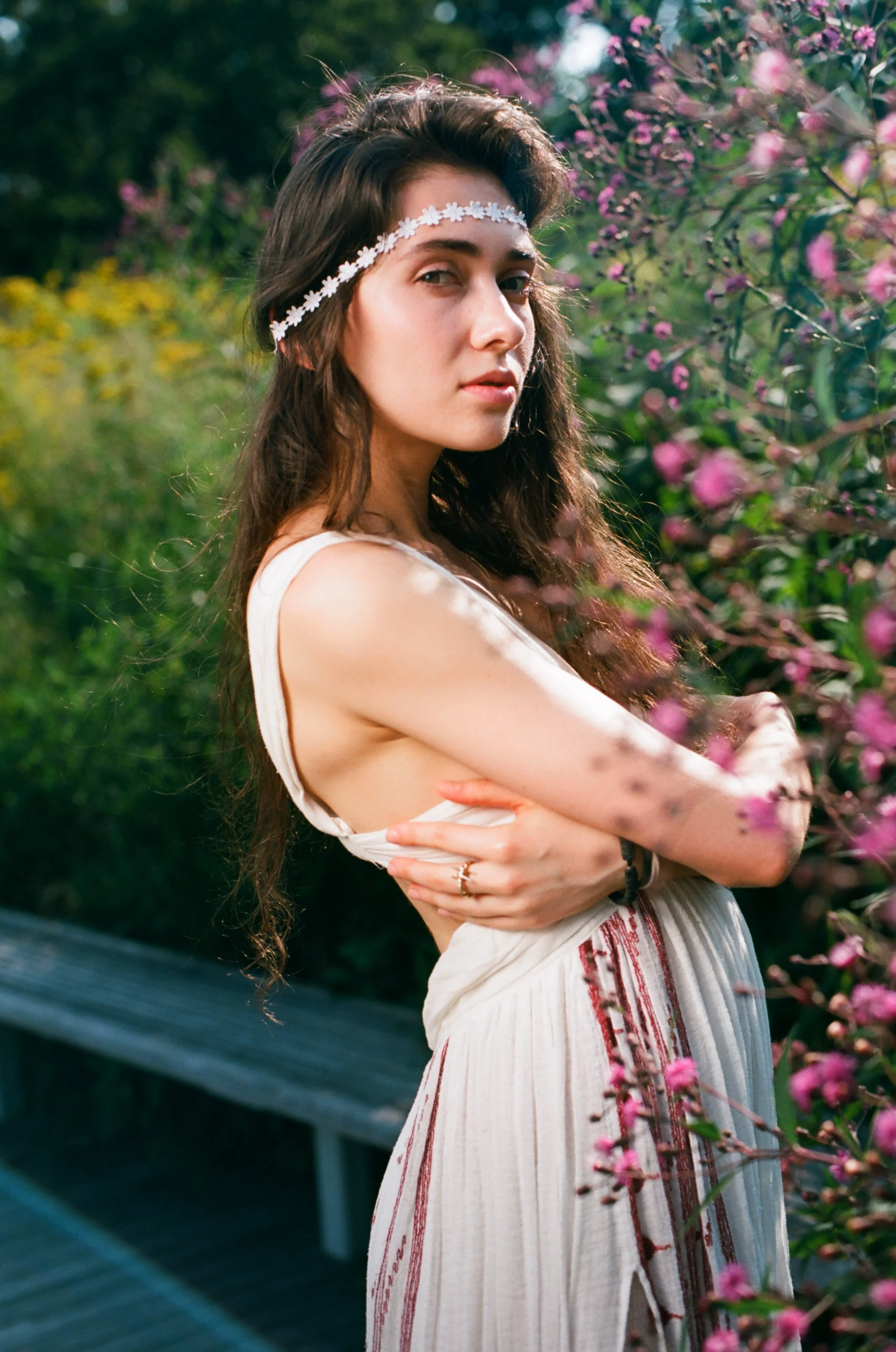 A young woman with long, wavy dark hair wearing a white flower headband and a sleeveless white dress with red embroidery, standing outdoors among pink flowering bushes. She has her arms crossed over her chest and is looking at the camera with a seren