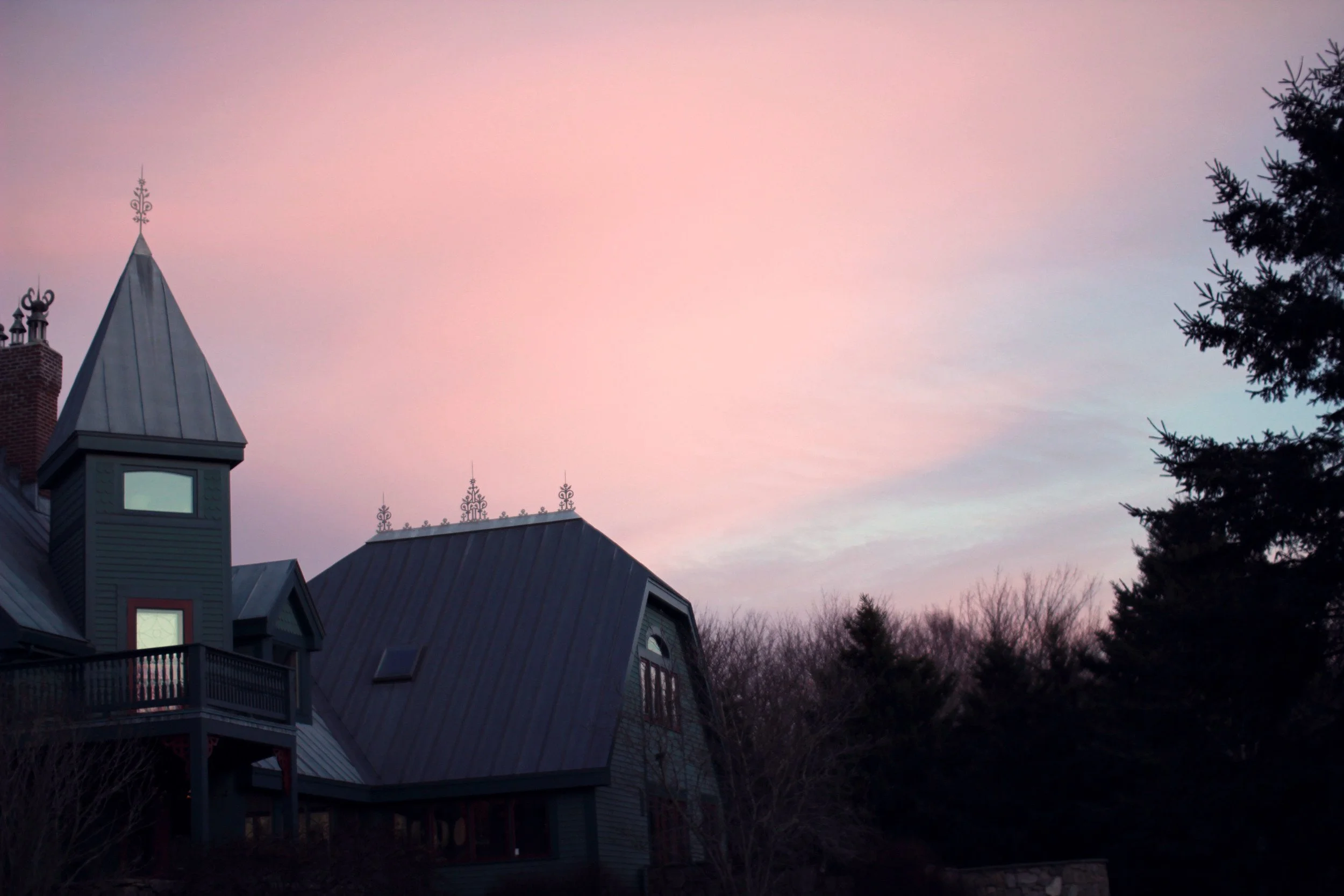 A dark, Victorian-style house with steep roofs, towers, and decorative ironwork on rooftops, silhouetted against a colorful sunset sky with pink and purple hues, surrounded by trees.