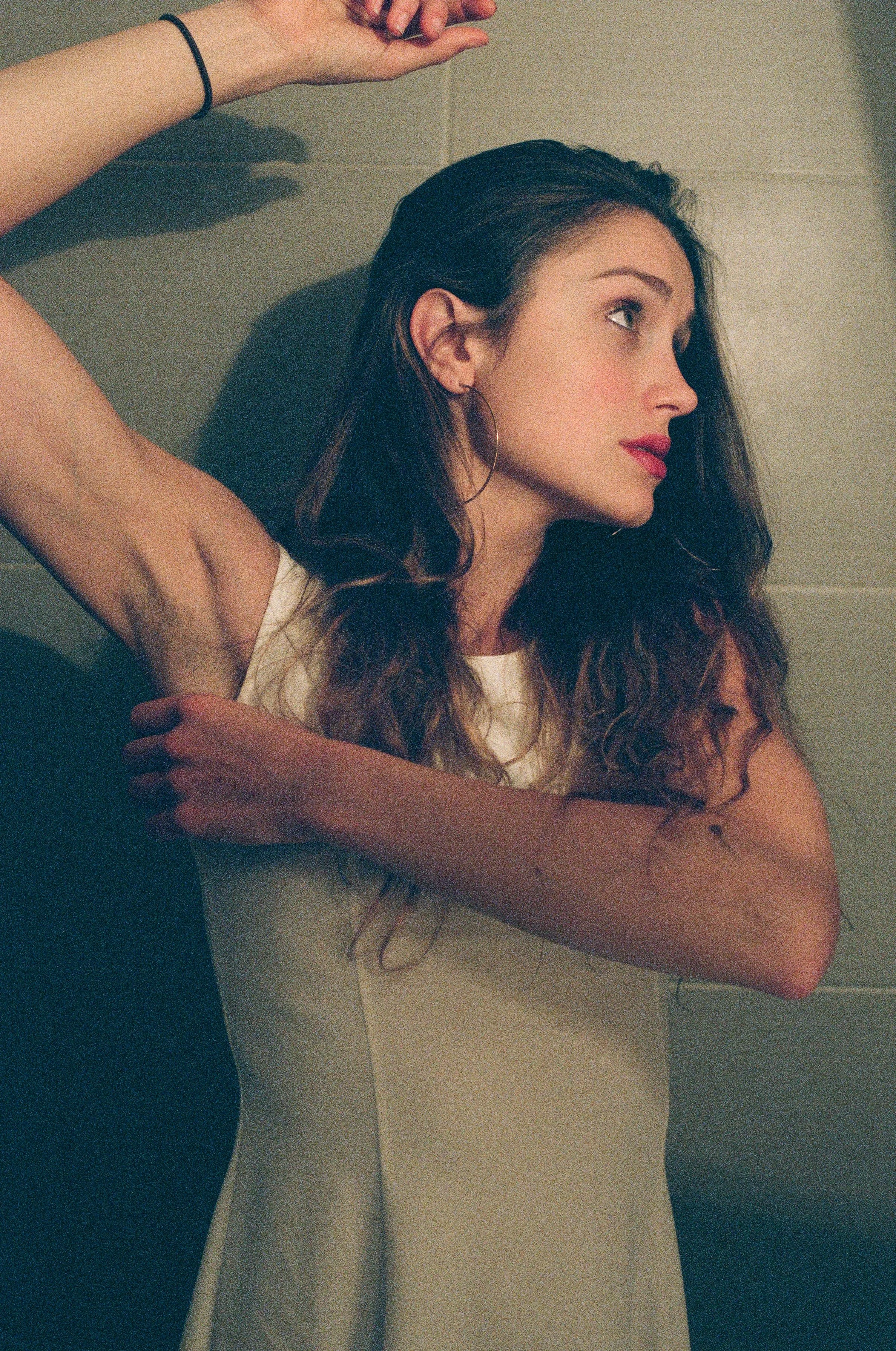 A young woman with long wavy brown hair, wearing hoop earrings and a sleeveless white top, lying on her side against a tiled wall, looking to her right with one arm raised above her head and the other across her chest.