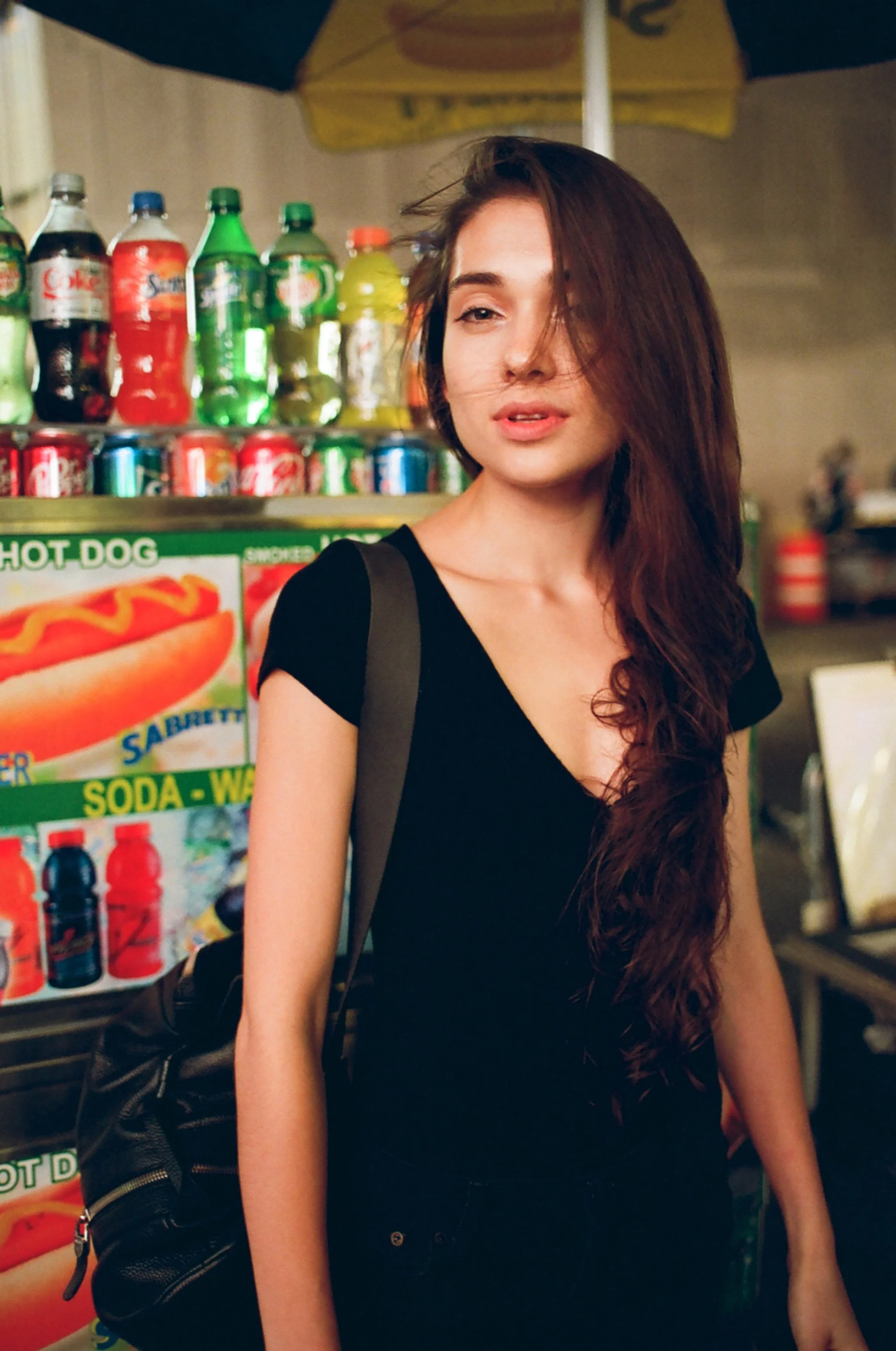 A young woman with long, wavy brown hair wearing a black top standing in front of a snack stand with various soda bottles and cans, and a hot dog sign.