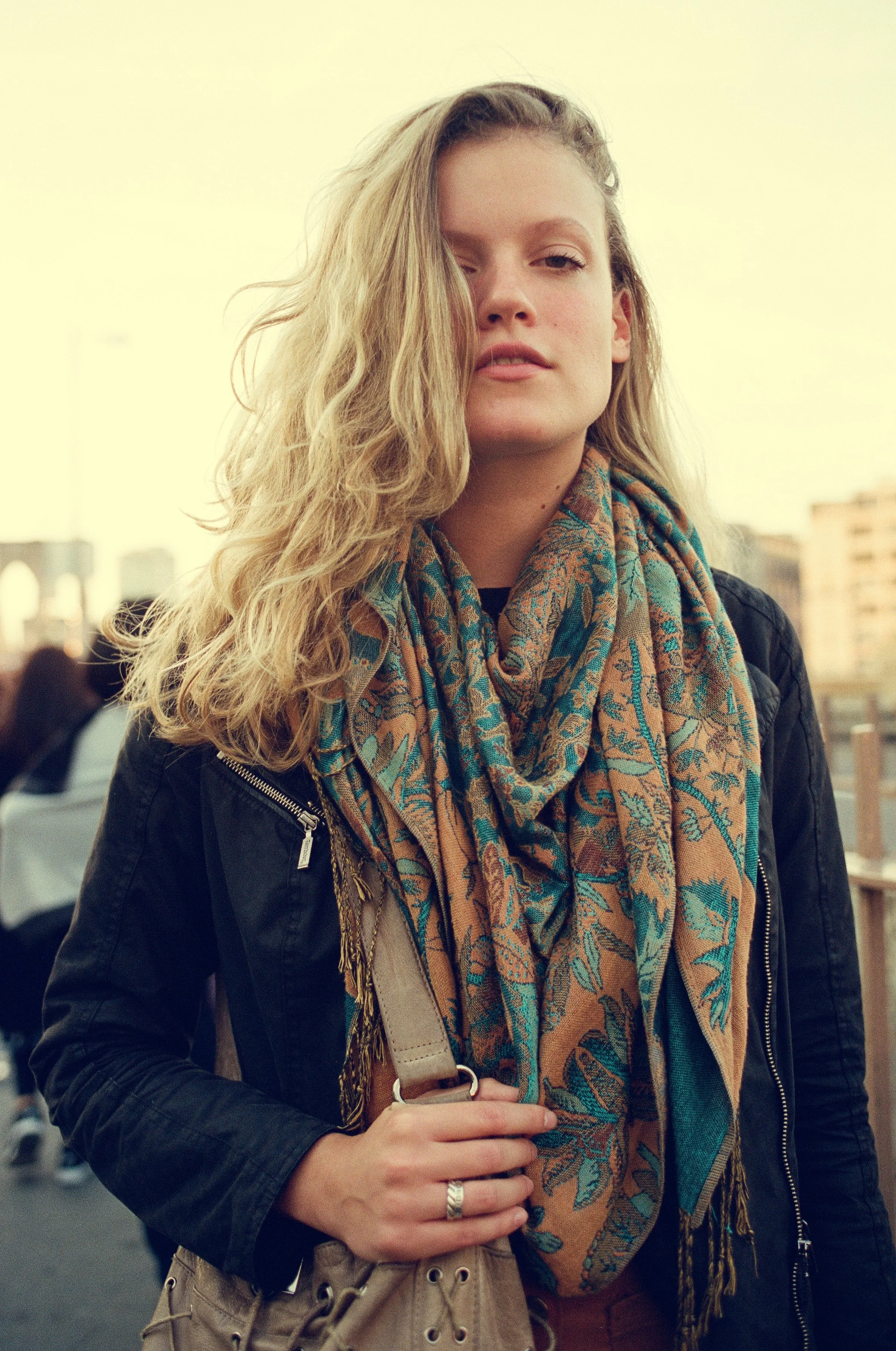 A young woman with wavy blonde hair wearing a black jacket and a colorful patterned scarf, standing outdoors in an urban setting.