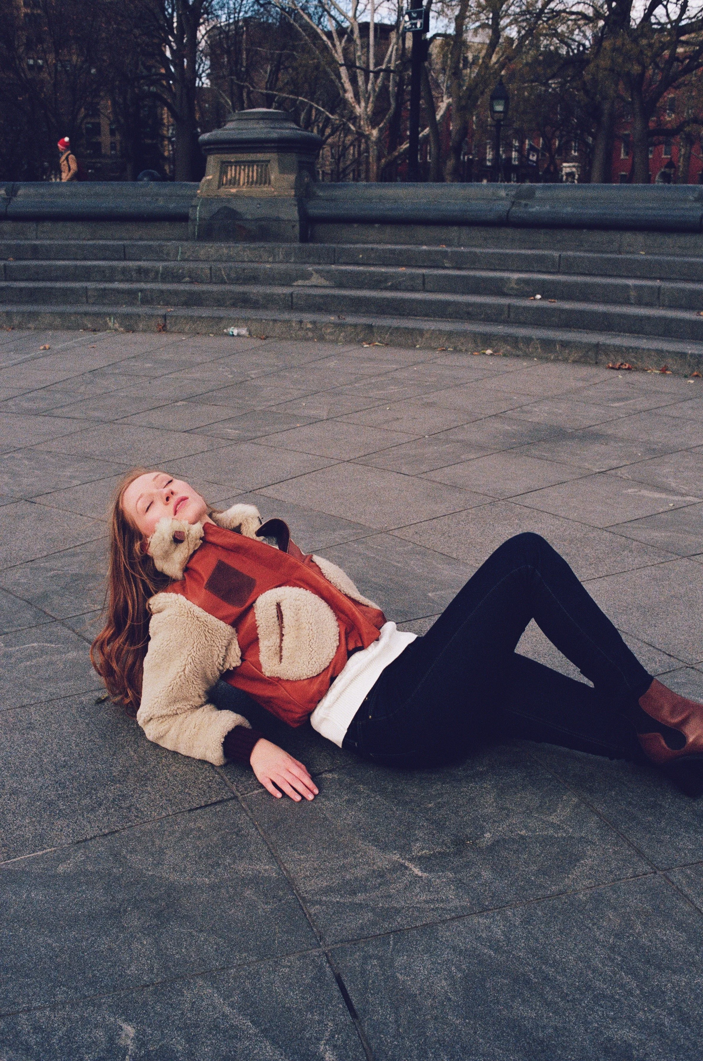 A woman with red hair lying on the pavement in a park, with her eyes closed and a relaxed expression, wearing a tan and red jacket, black pants, and brown boots.